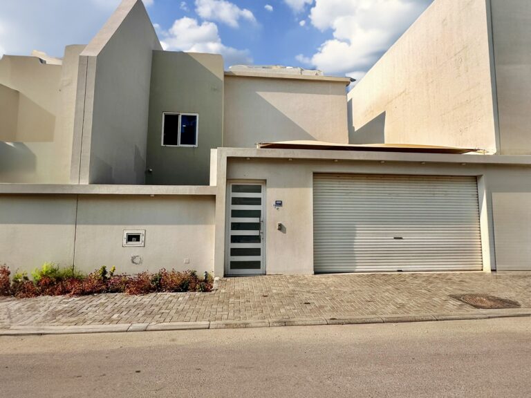 A modern, minimalist beige house with a flat roof, featuring a large garage door and a small window on the upper left side. The house is situated on a paved street with a patch of shrubs to the left, giving it an auto draft-like precision and clean aesthetic.