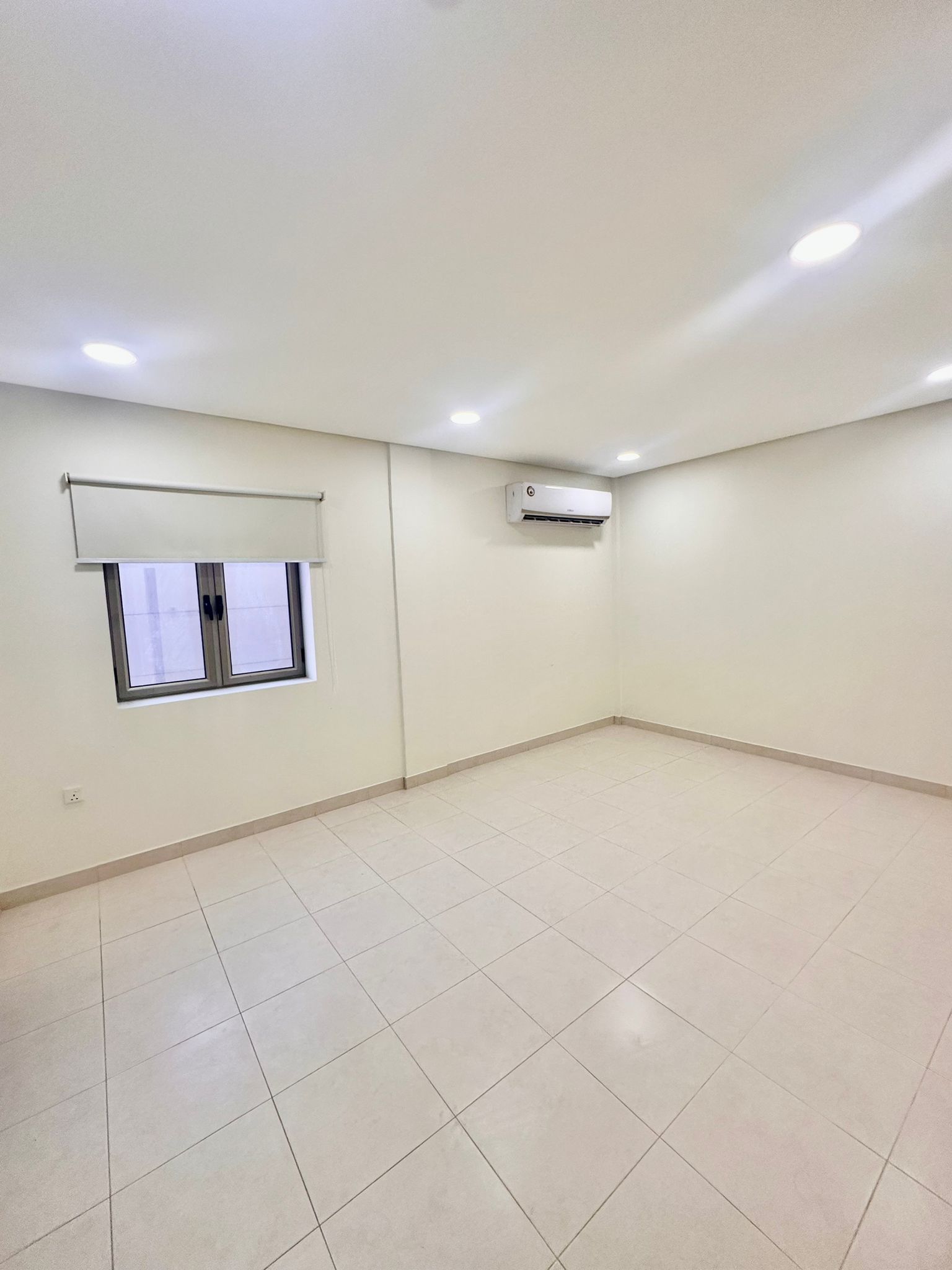 A clean, spacious room with white walls, tiled floor, a window with a roller blind, an air conditioning unit on the wall, and recessed ceiling lights in the Shakurah Triplex Apartment.