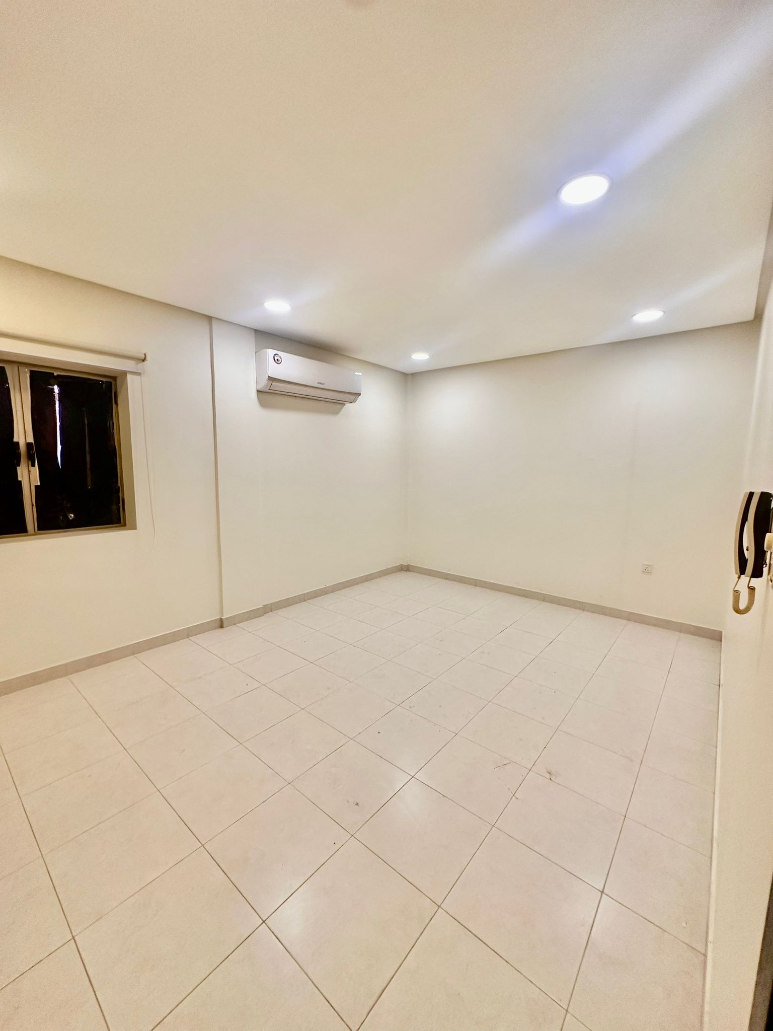 An empty room with white tiled flooring, white walls, a window with vertical blinds, an air conditioner mounted on the wall, and ceiling lights turned on. This spacious area in the Shakurah Triplex Apartment is both modern and inviting.