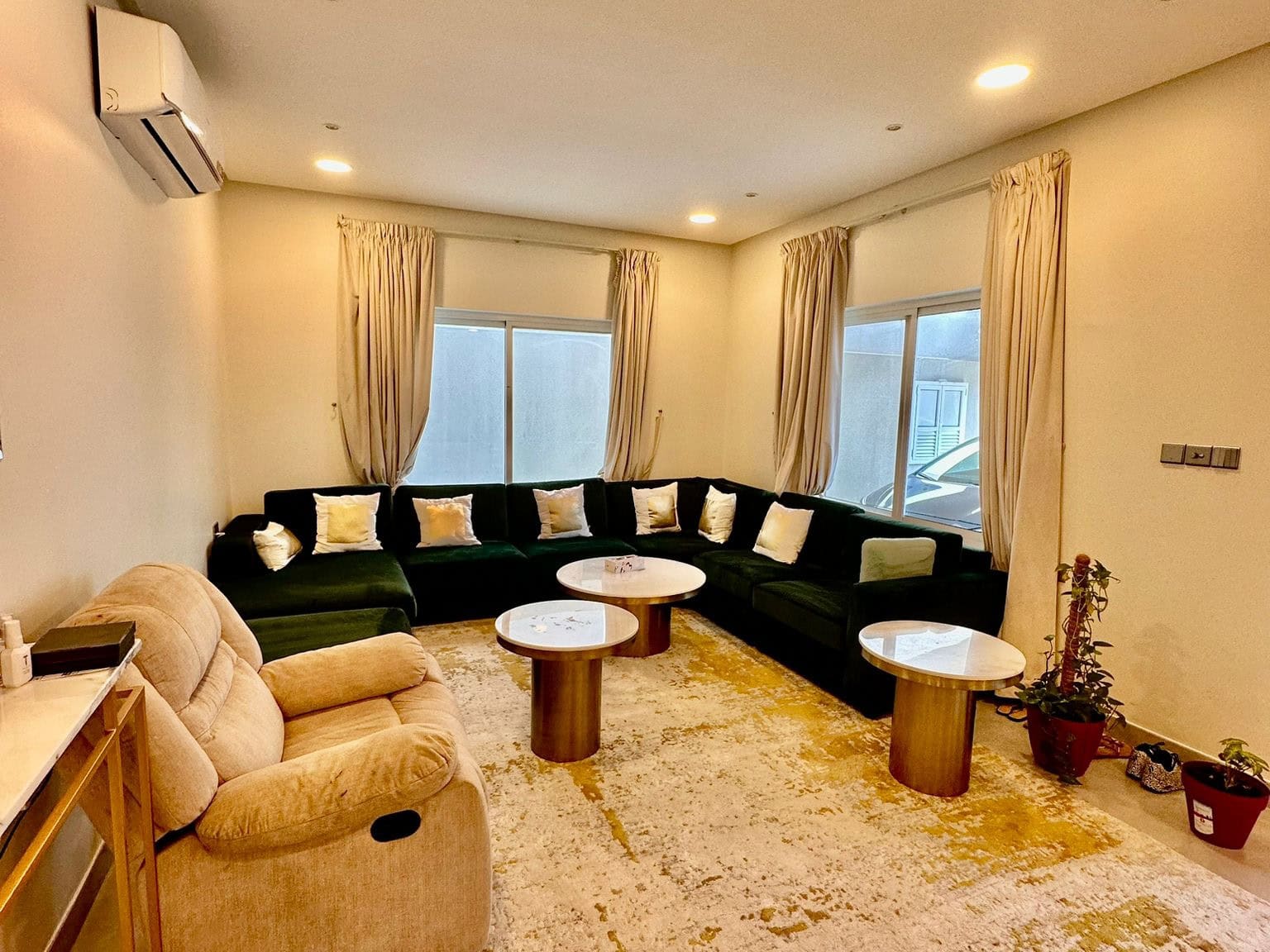 A cozy living room with a large L-shaped dark green sofa, beige recliner, two round white tables, and curtains. The space features an air conditioner and several windows with outdoor views, offering a refreshing escape from the daily grind. It's the perfect setting for some much-needed auto draft relaxation.