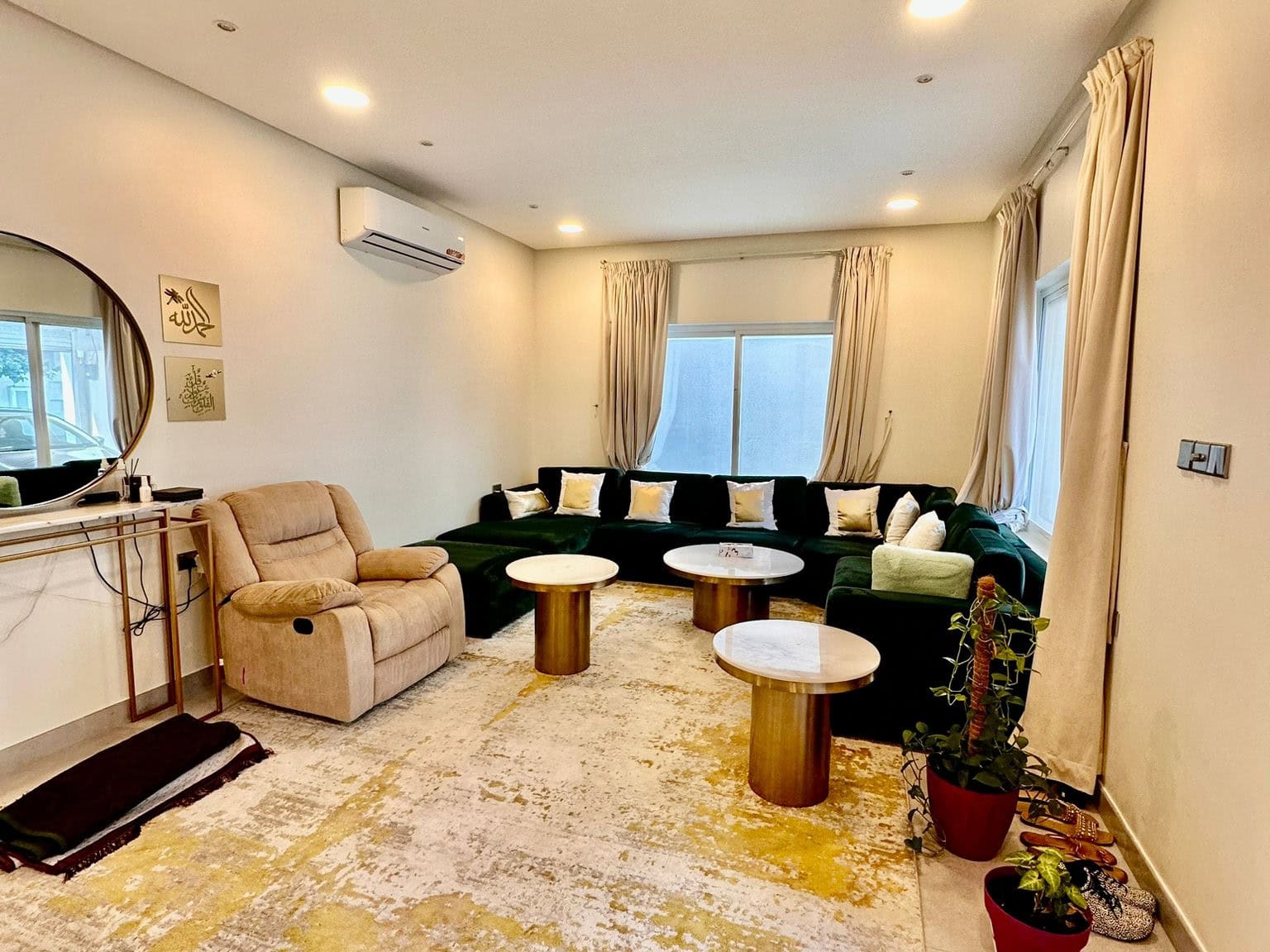 A cozy living room in a private villa in the Saar Area features a green L-shaped sofa, recliner chair, three round coffee tables, wall-mounted air conditioner, mirror, and large windows with cream curtains.