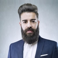 Man with a full beard and styled hair wearing a blue suit and white shirt, looking at the camera against a plain background.