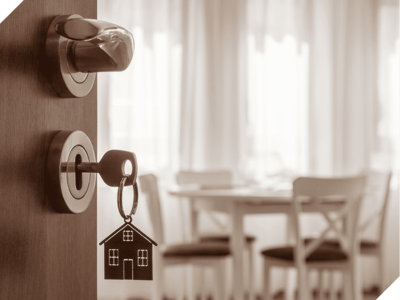 A key with a house-shaped keychain is inserted in a door lock; a blurred interior with chairs and a table hints at property paperwork awaiting the real estate agents inside.