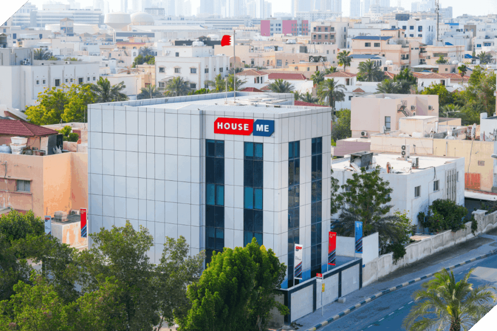 A modern, white commercial building labeled "House ME" stands among residential houses in Bahrain, with a red and white flag on the roof—showcasing House Me Bahrain company’s expert commercial real estate services against a backdrop of city buildings.