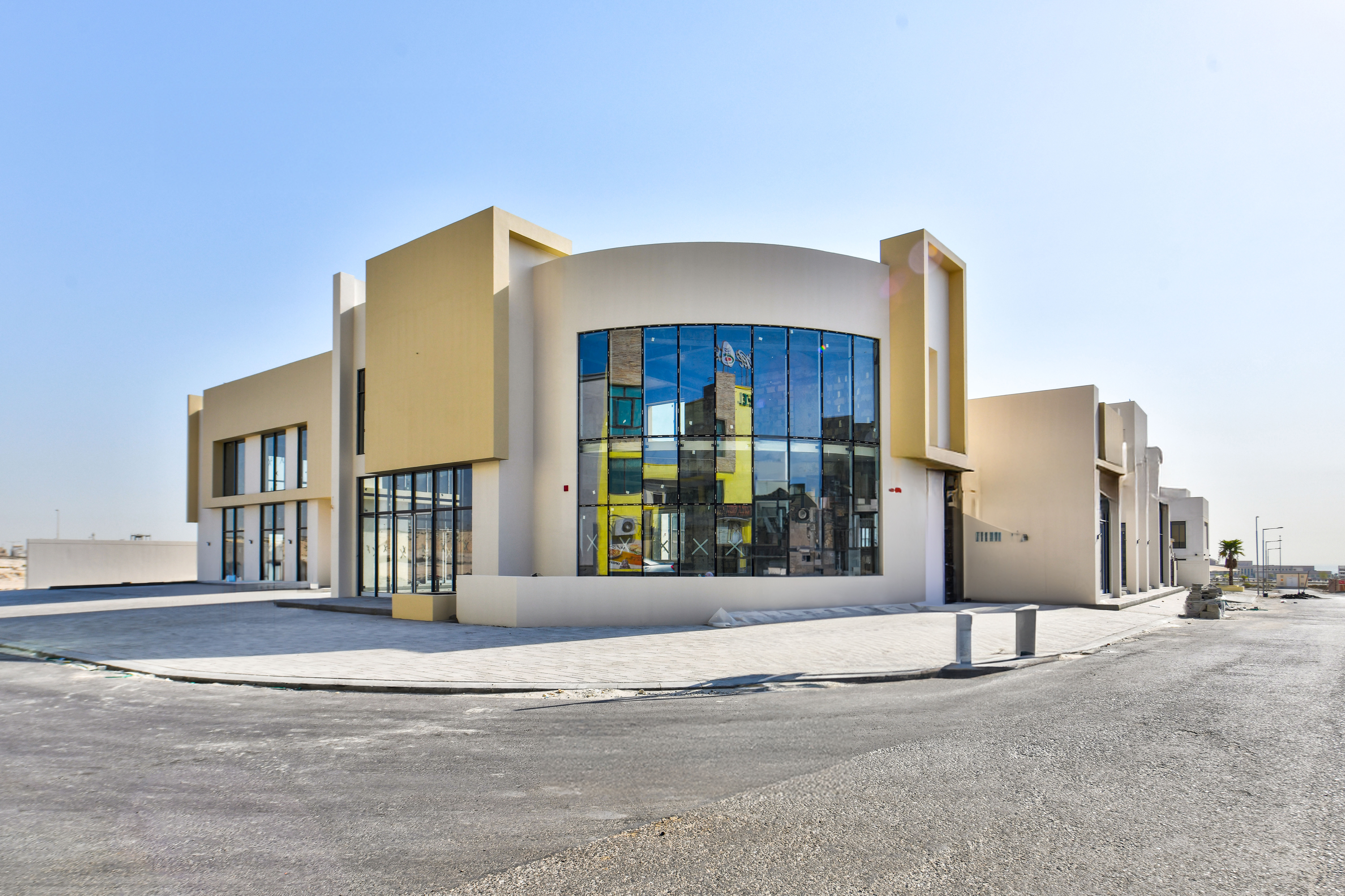 Modern commercial building with large reflective glass windows and a beige exterior, situated on a paved corner lot in Askar Square under a clear blue sky.
