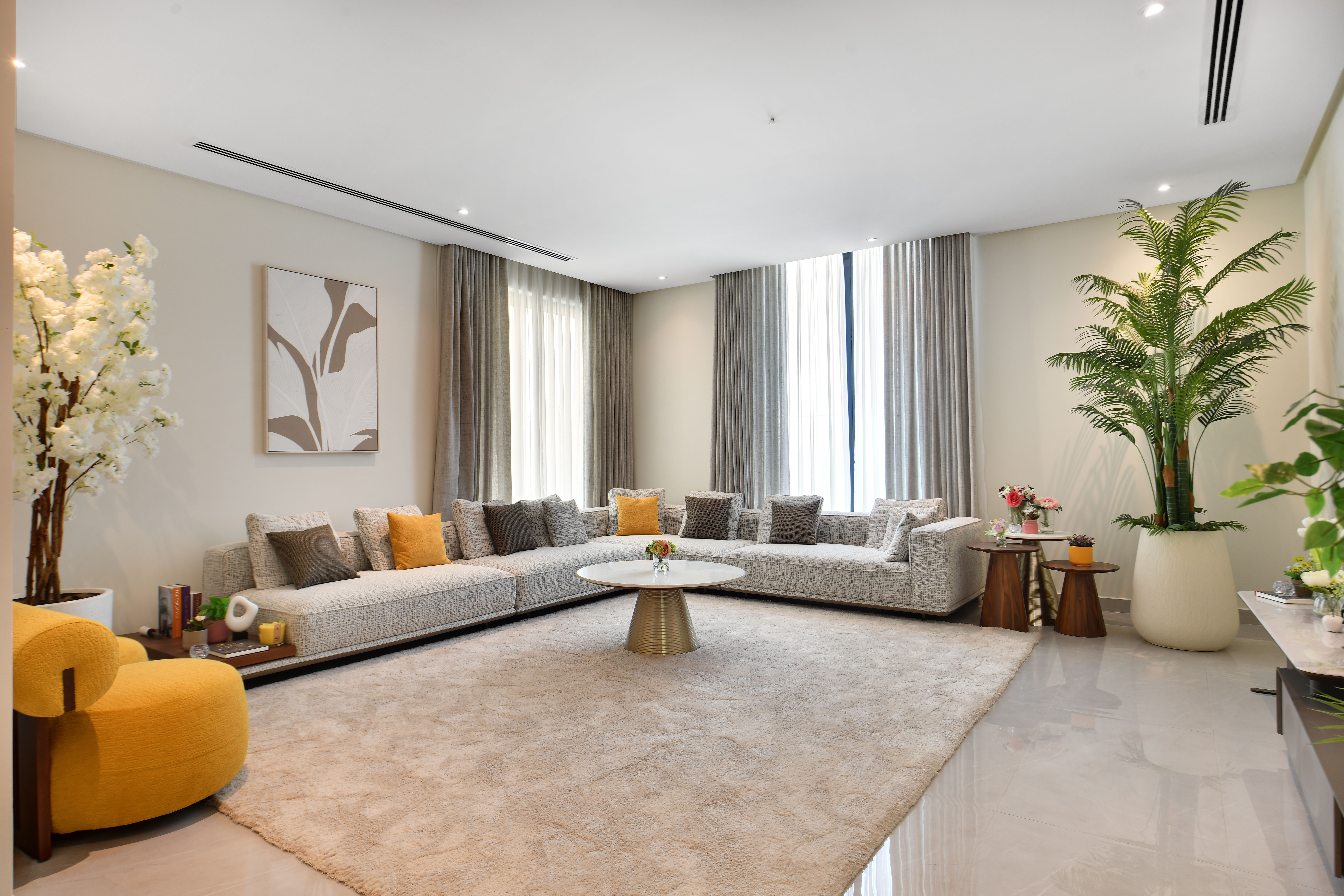 Modern living room at Durrah Villas with a large sectional sofa, round coffee table, beige rug, yellow armchair, potted plants, and abstract wall art, all illuminated by natural light from tall windows.