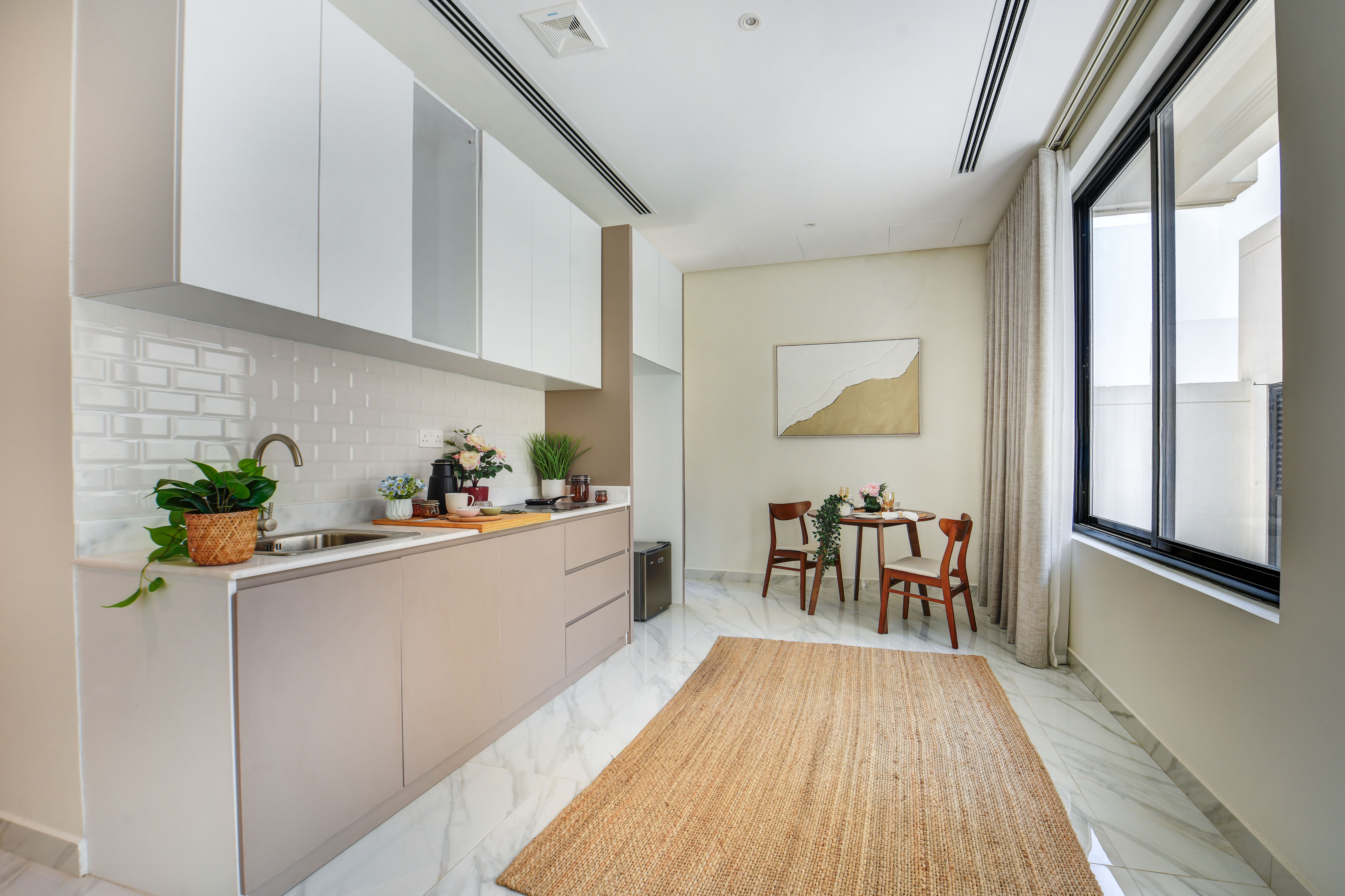 Modern kitchen at Durrah Villas with light cabinets, white tile backsplash, indoor plants, a small dining table for two, large window, and a woven rug on a marble floor.