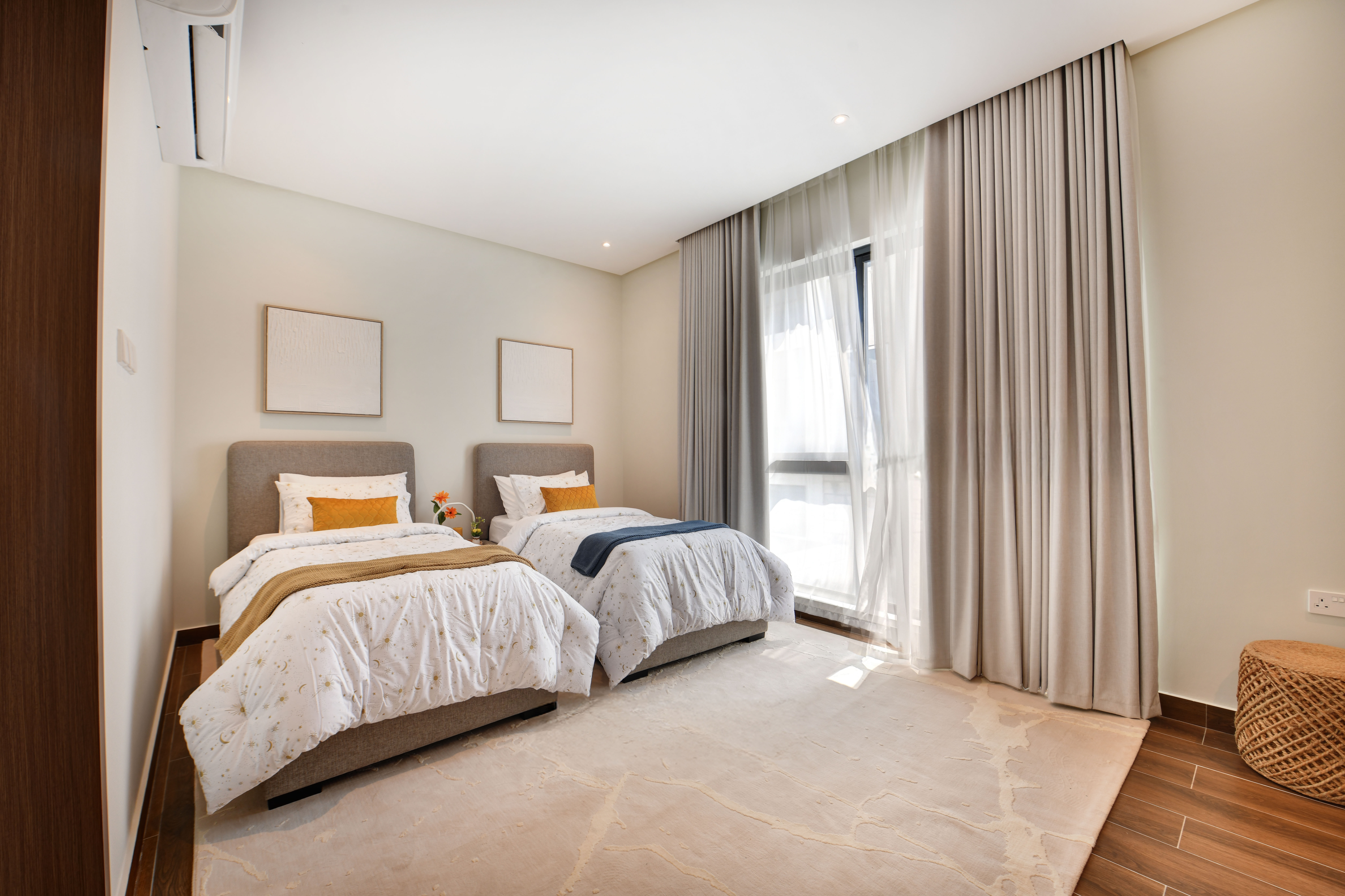 Modern bedroom at Durrah Villas with two single beds, white bedding, a large window with sheer curtains, wooden floor, two wall frames, and a round woven ottoman.