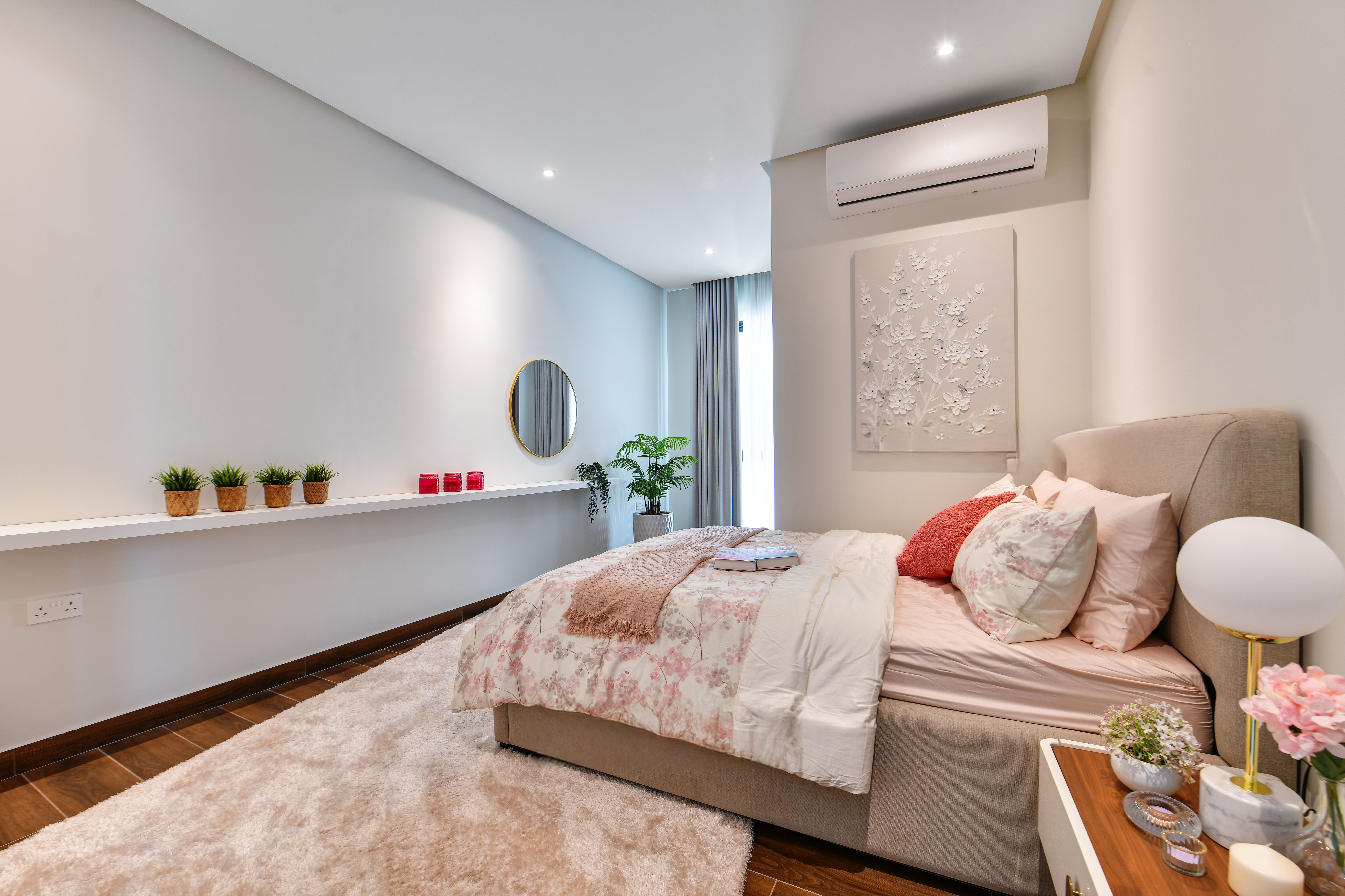 A neatly made bed with floral bedding in a modern Durrah Villas bedroom, featuring wall art, potted plants, a round mirror, and a white rug on a wooden floor.