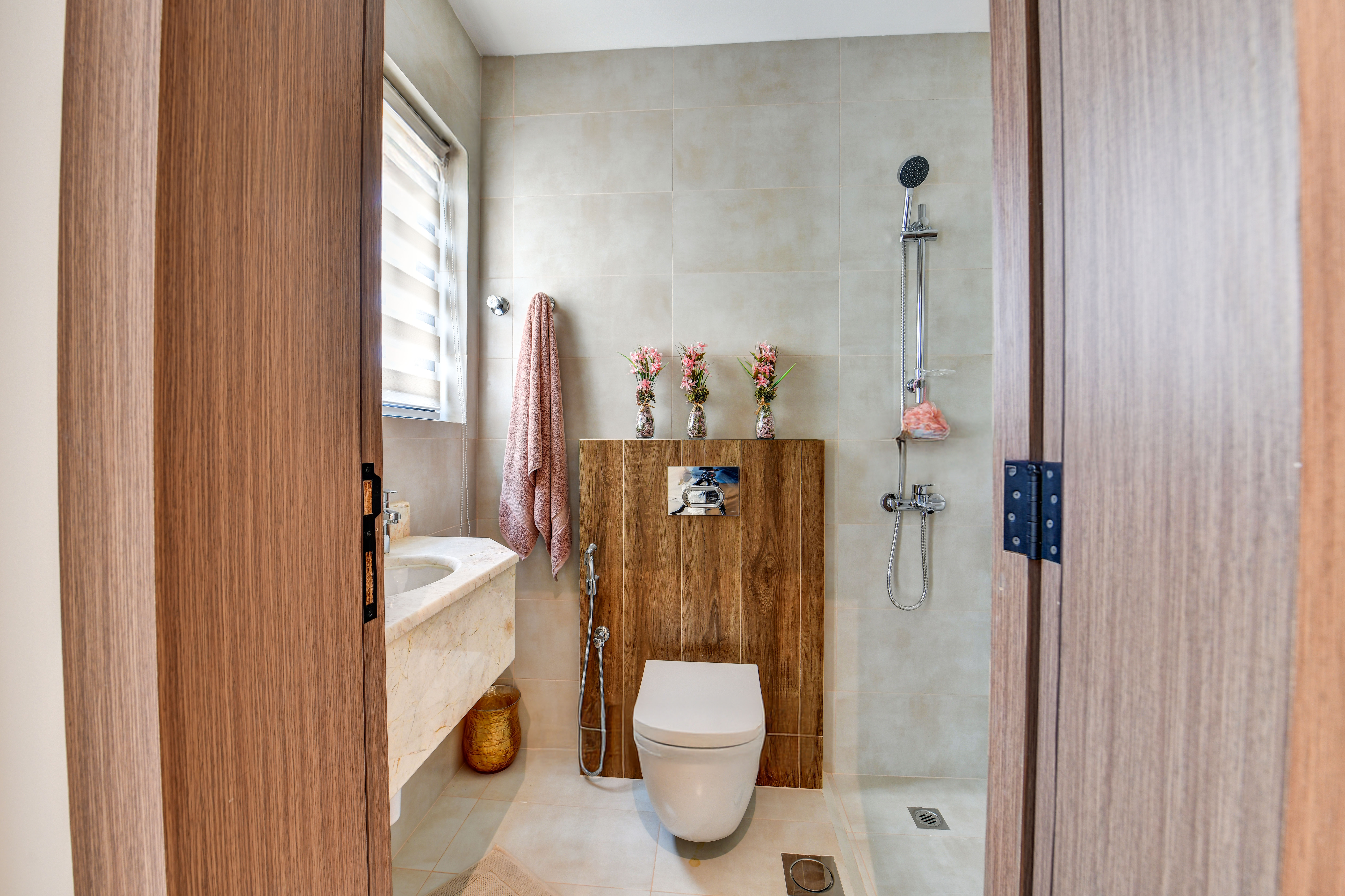 Modern bathroom at Durrah Villas with a wall-mounted toilet, shower, sink, striped window blind, and three vases with flowers on a wooden shelf above the toilet.