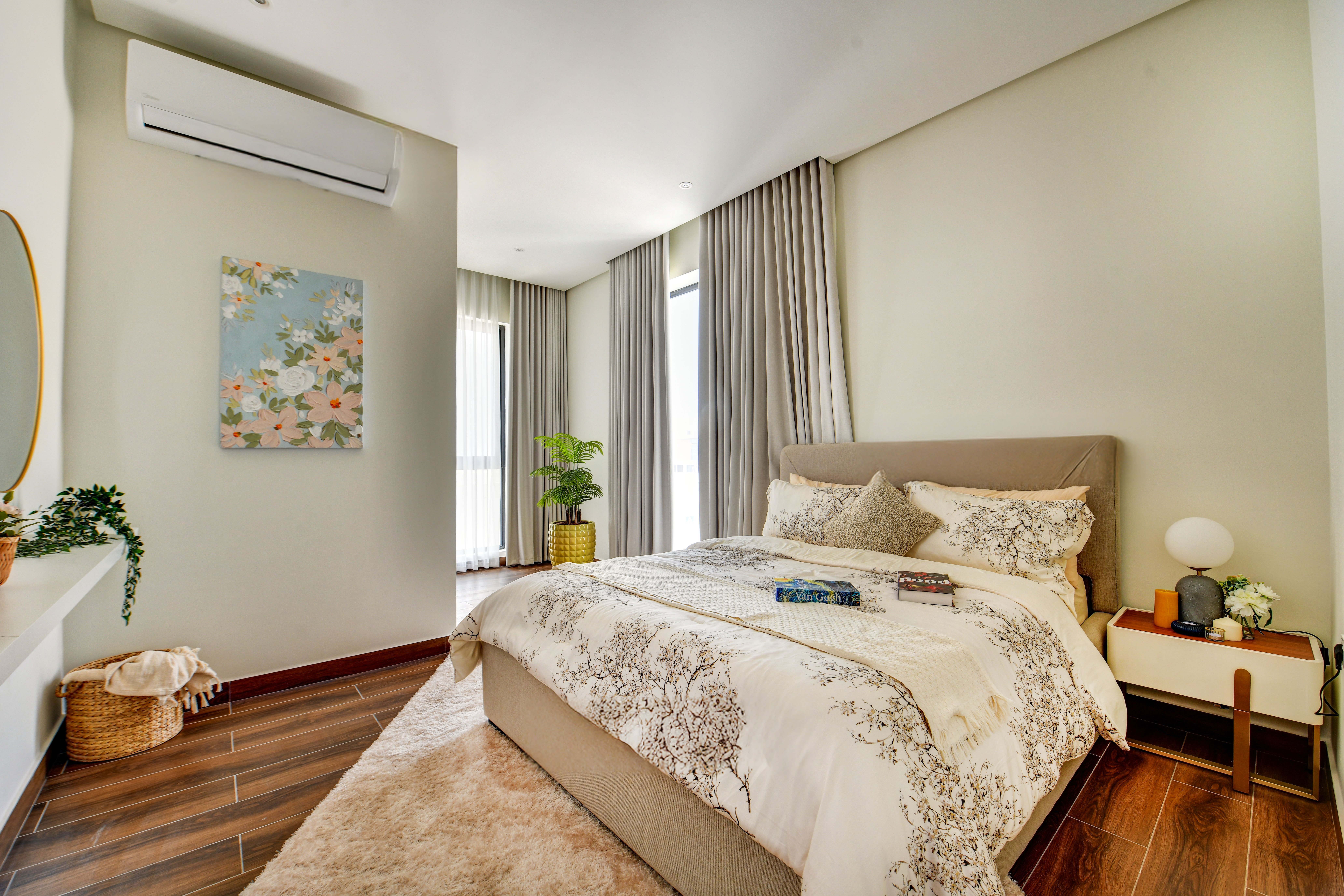 A modern bedroom at Durrah Villas features a large bed, neutral bedding, nightstand, plants, wall art, wooden flooring, and sunlight streaming through tall windows with sheer curtains.