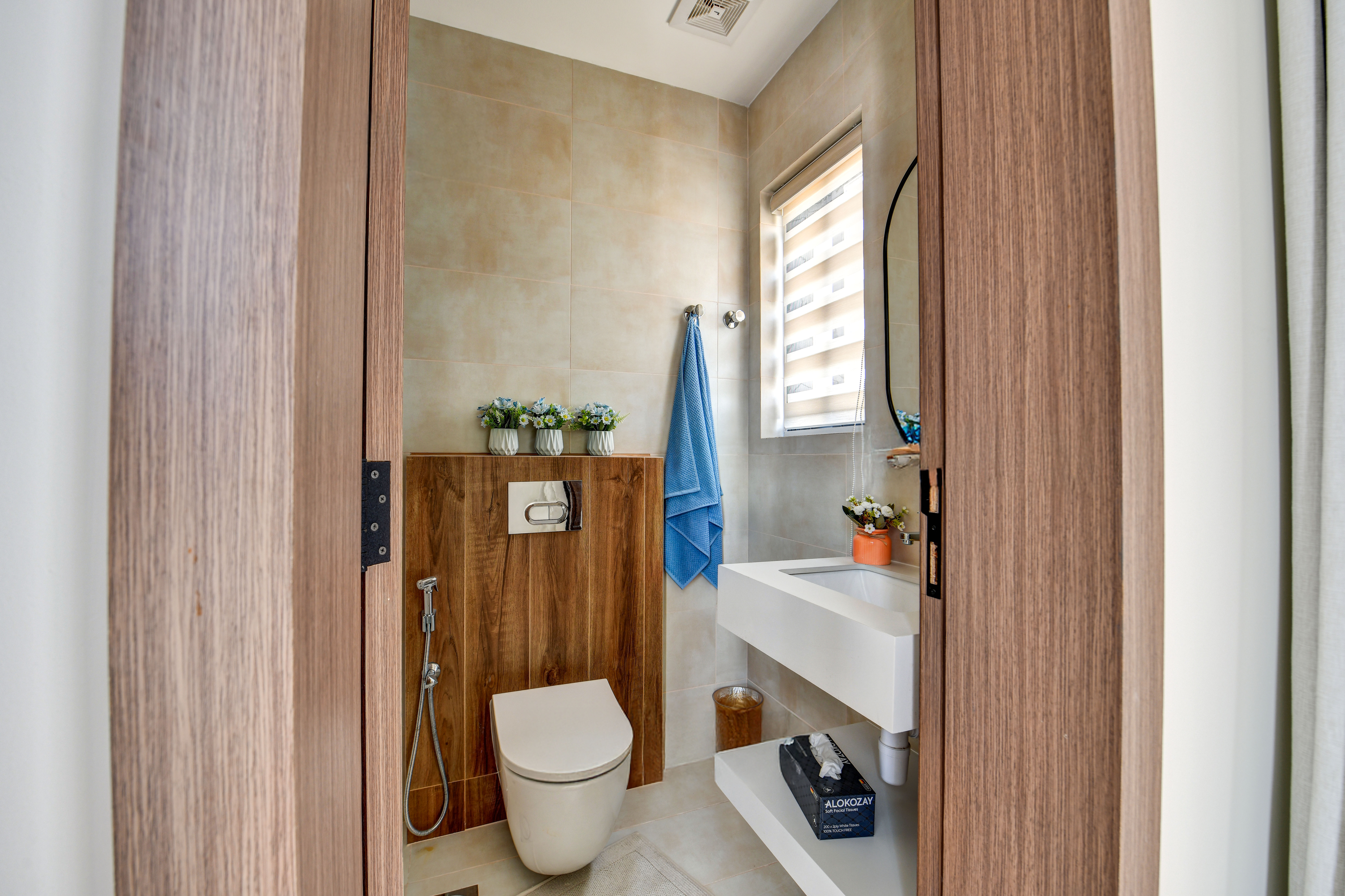 Small modern bathroom at Durrah Villas with a wall-mounted toilet, wooden accents, a white sink, blue towel, decorative flowers, and a window with blinds.