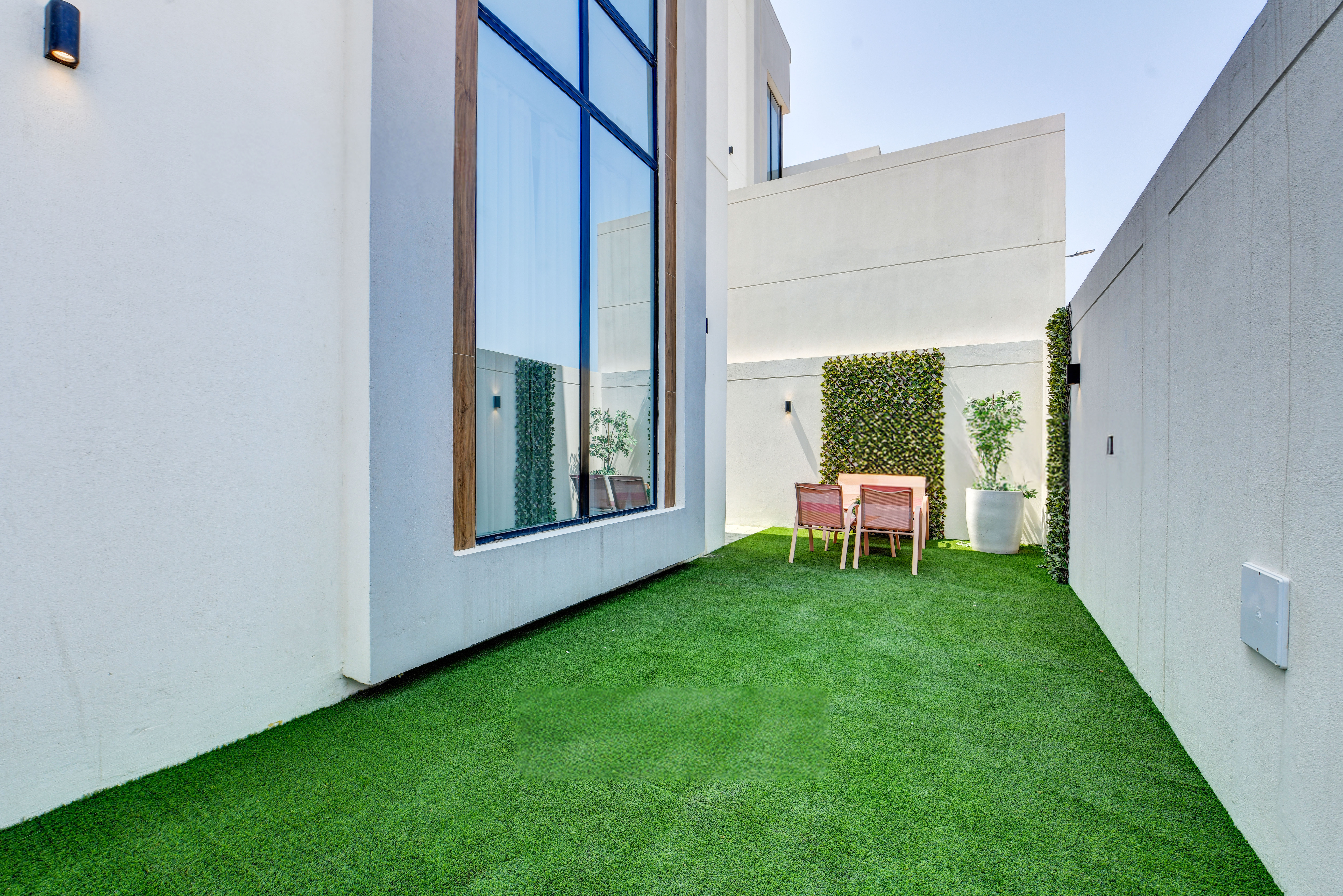 A modern outdoor area at Durrah Villas with artificial grass, a dining table for four, tall windows, and white walls adorned with vertical green plants.