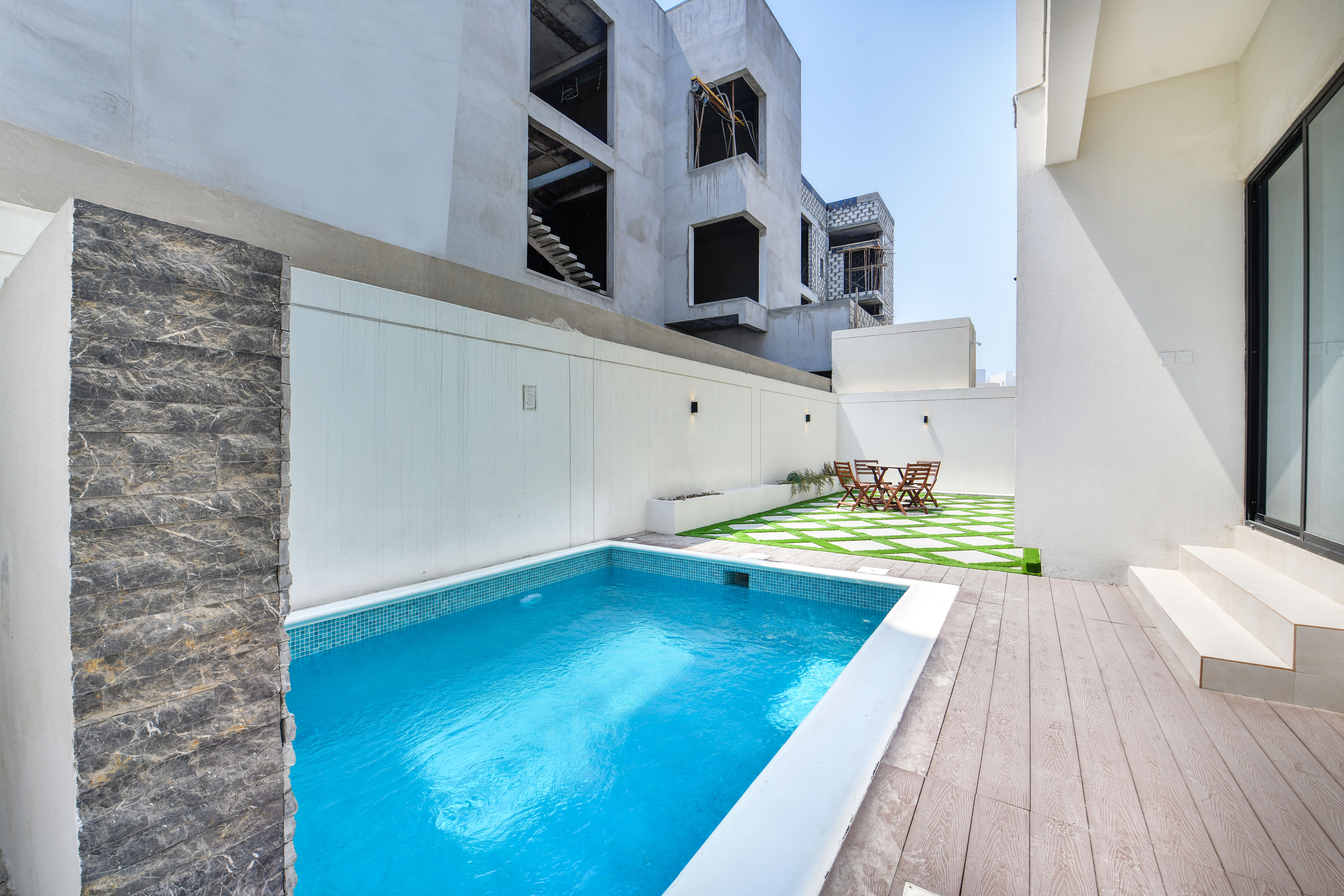 A small backyard at Durrah Villas with a rectangular swimming pool, stone water feature, wooden deck, and a grassy area with table and chairs, all enclosed by white walls.