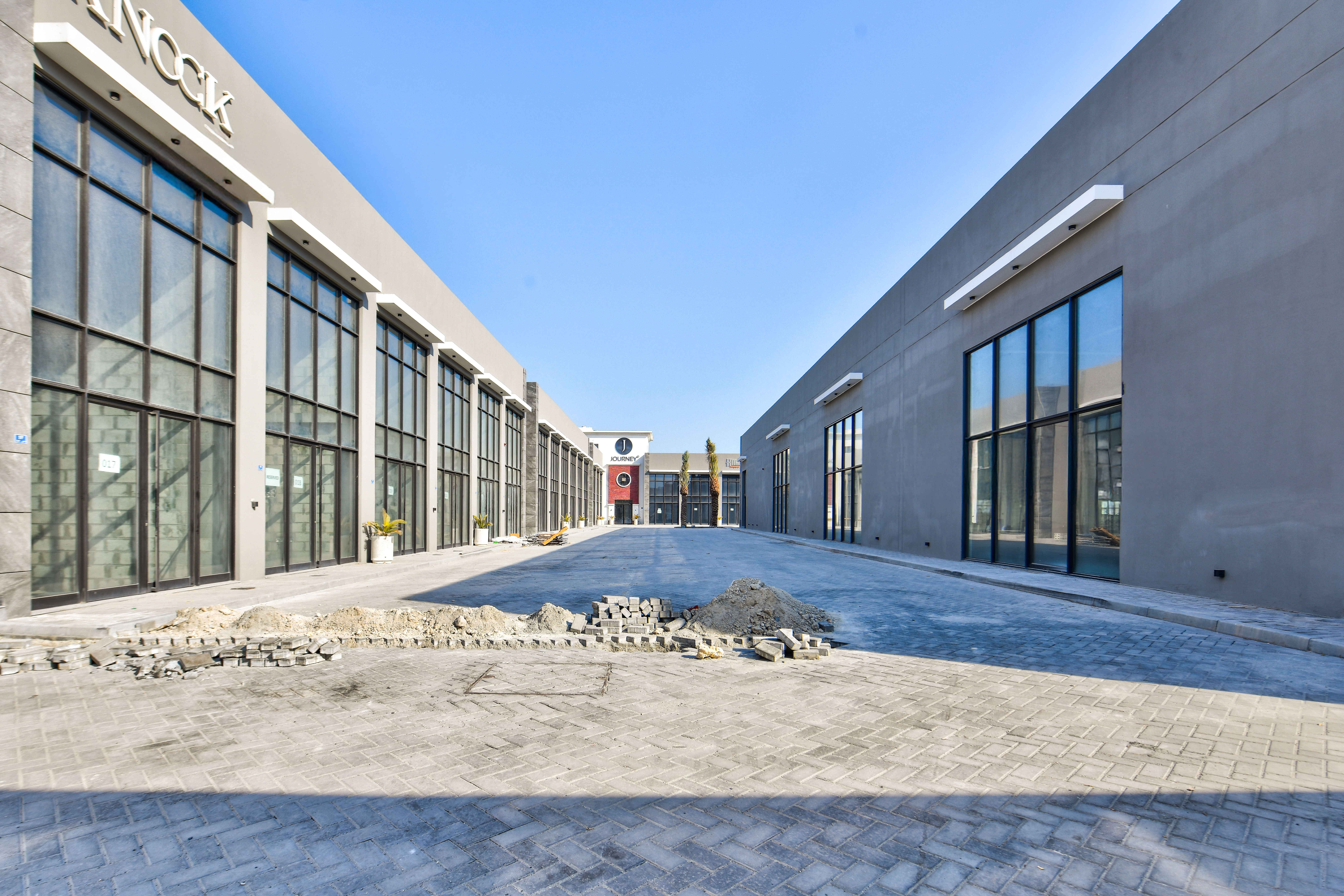 A wide, paved walkway at Sanad Square stretches between two modern commercial buildings with glass windows, featuring construction materials and unfinished pavement in the center.