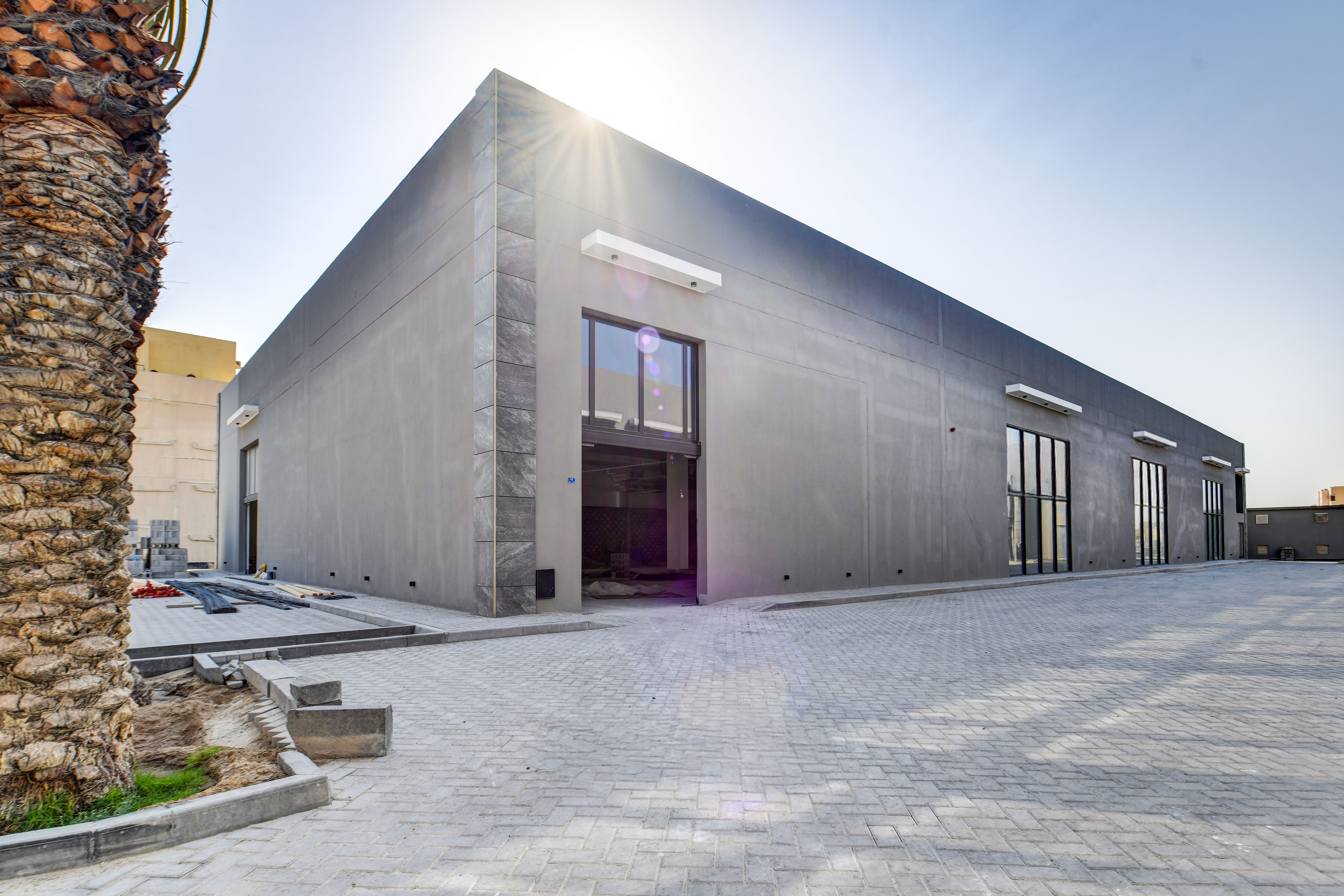 A modern, rectangular industrial building with large windows and a wide entrance, situated on a paved lot in Sanad Square under a clear sky.
