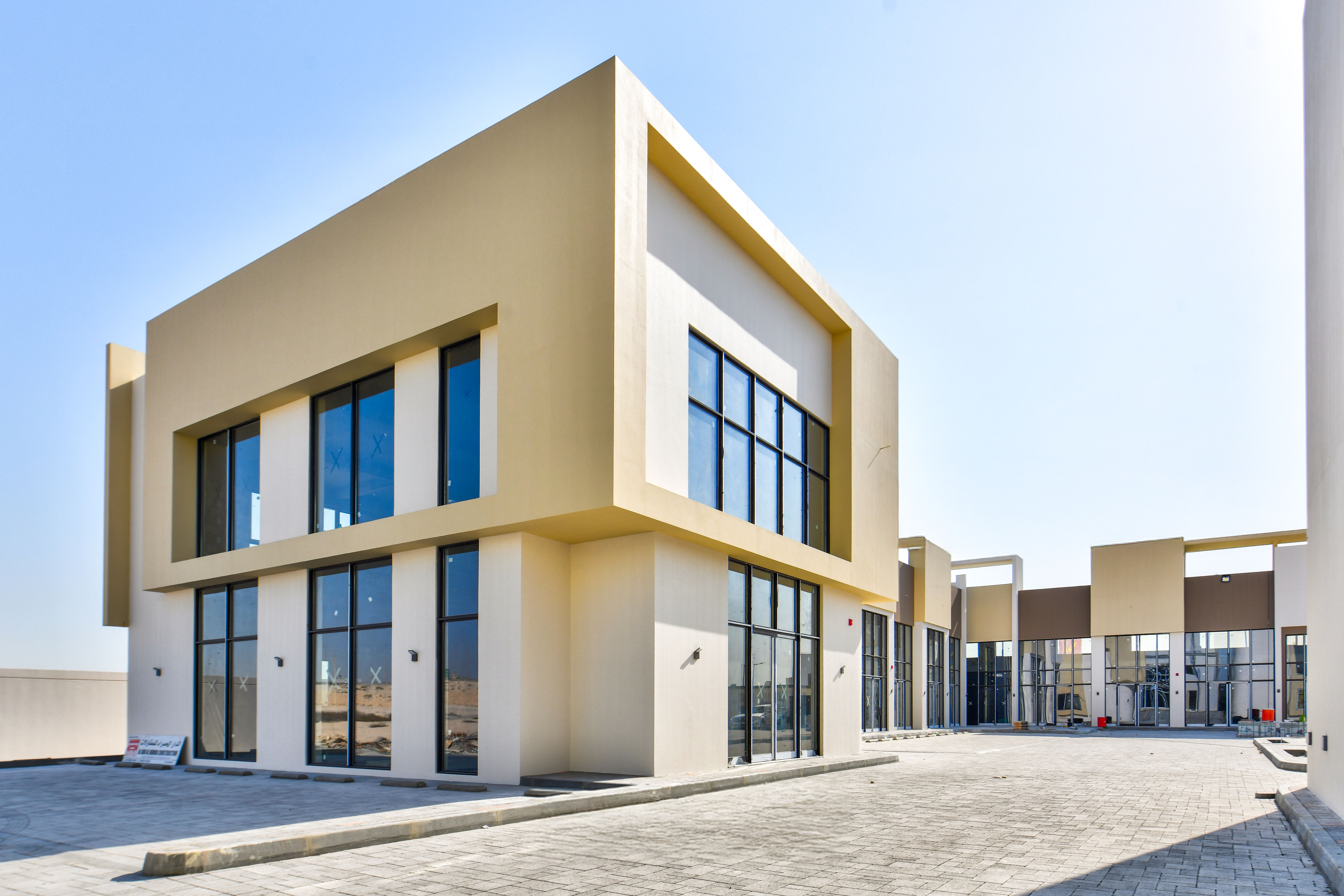 Modern two-story building with large glass windows and a beige exterior, situated along a paved road near Askar Square under a clear blue sky.