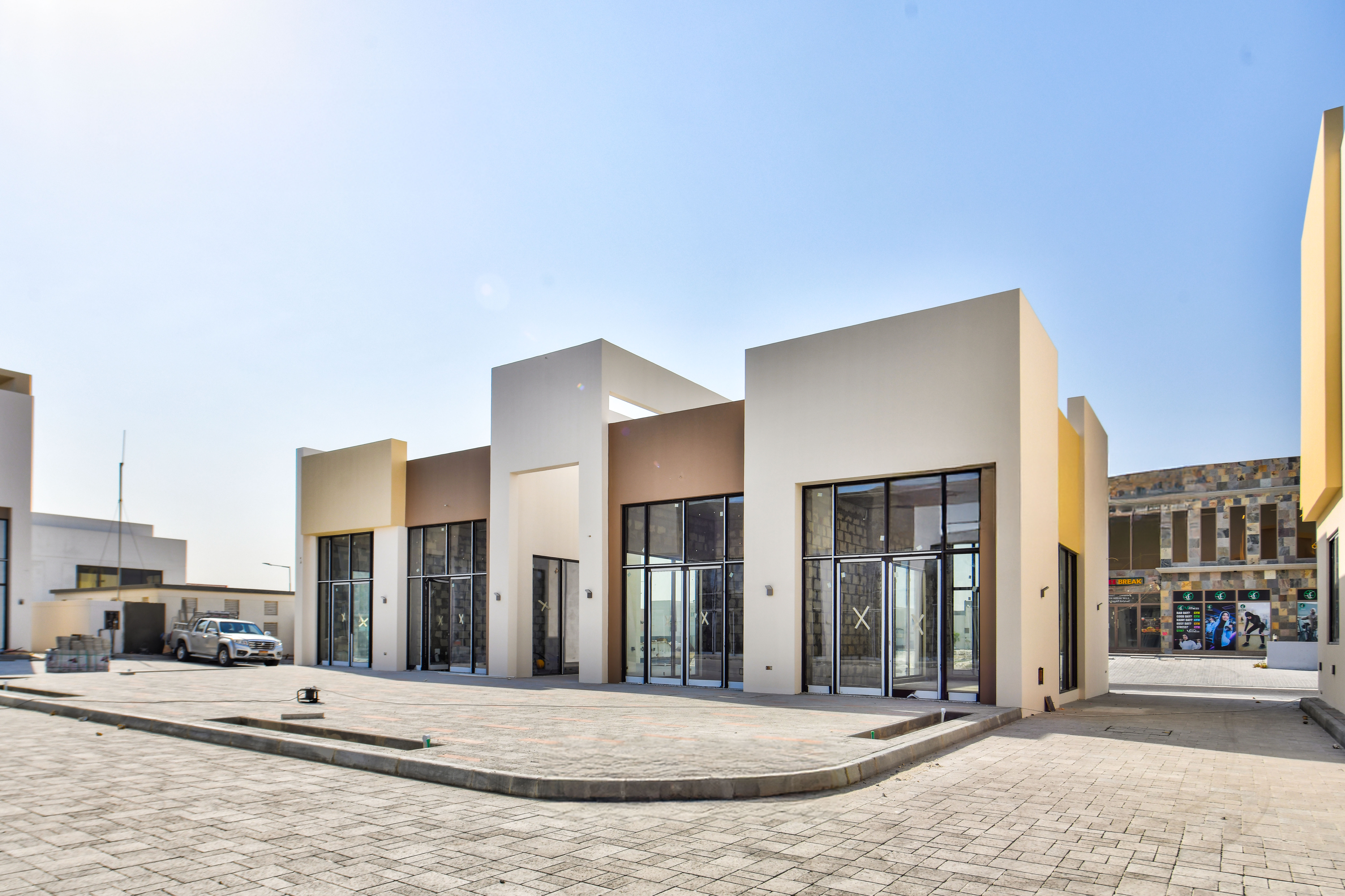 A modern, single-story commercial building with large glass windows stands near Askar Square, its paved area in front basking under a clear blue sky.