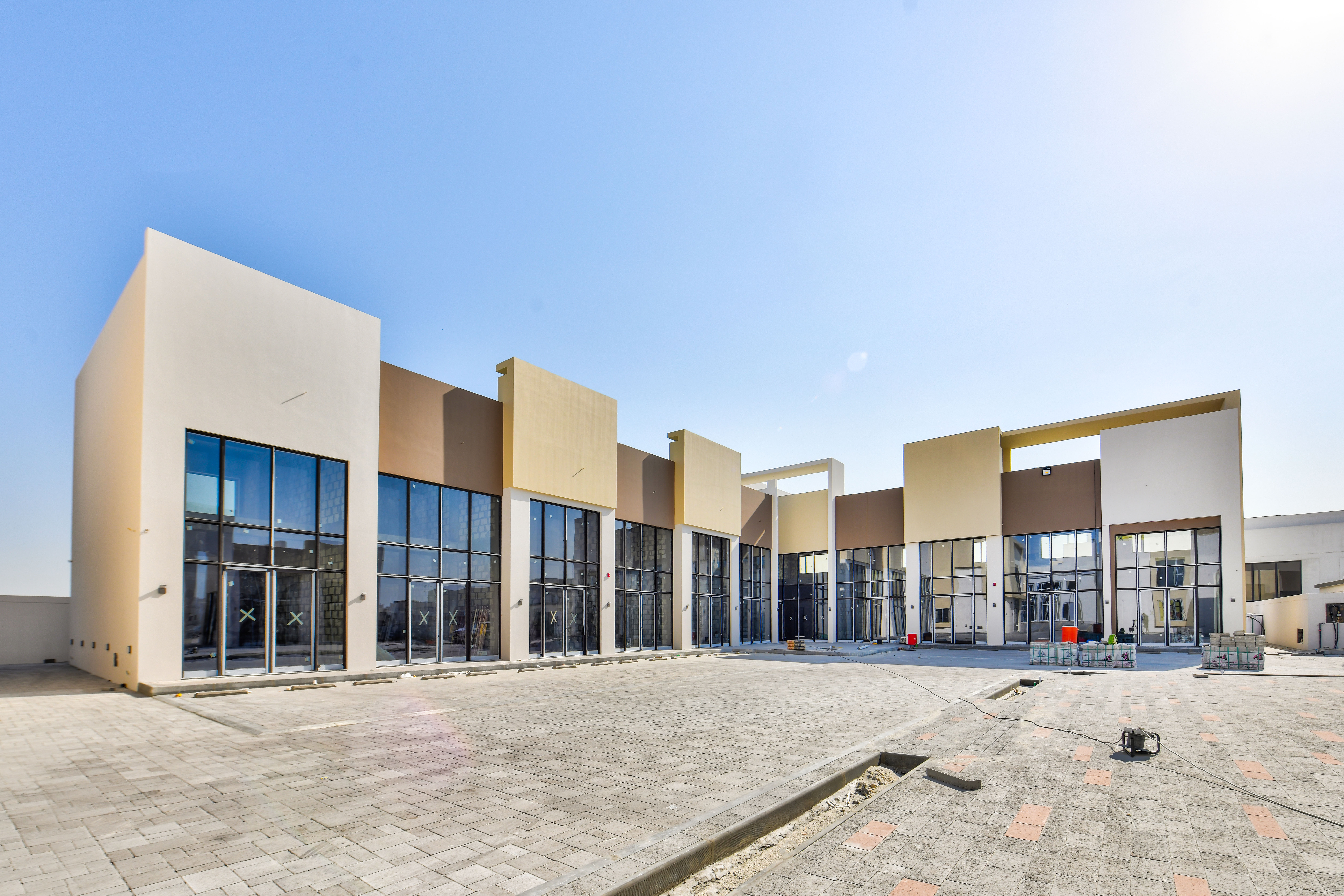 Modern commercial building with large glass windows, beige and brown exterior panels, and an empty paved courtyard at Askar Square under a clear blue sky.