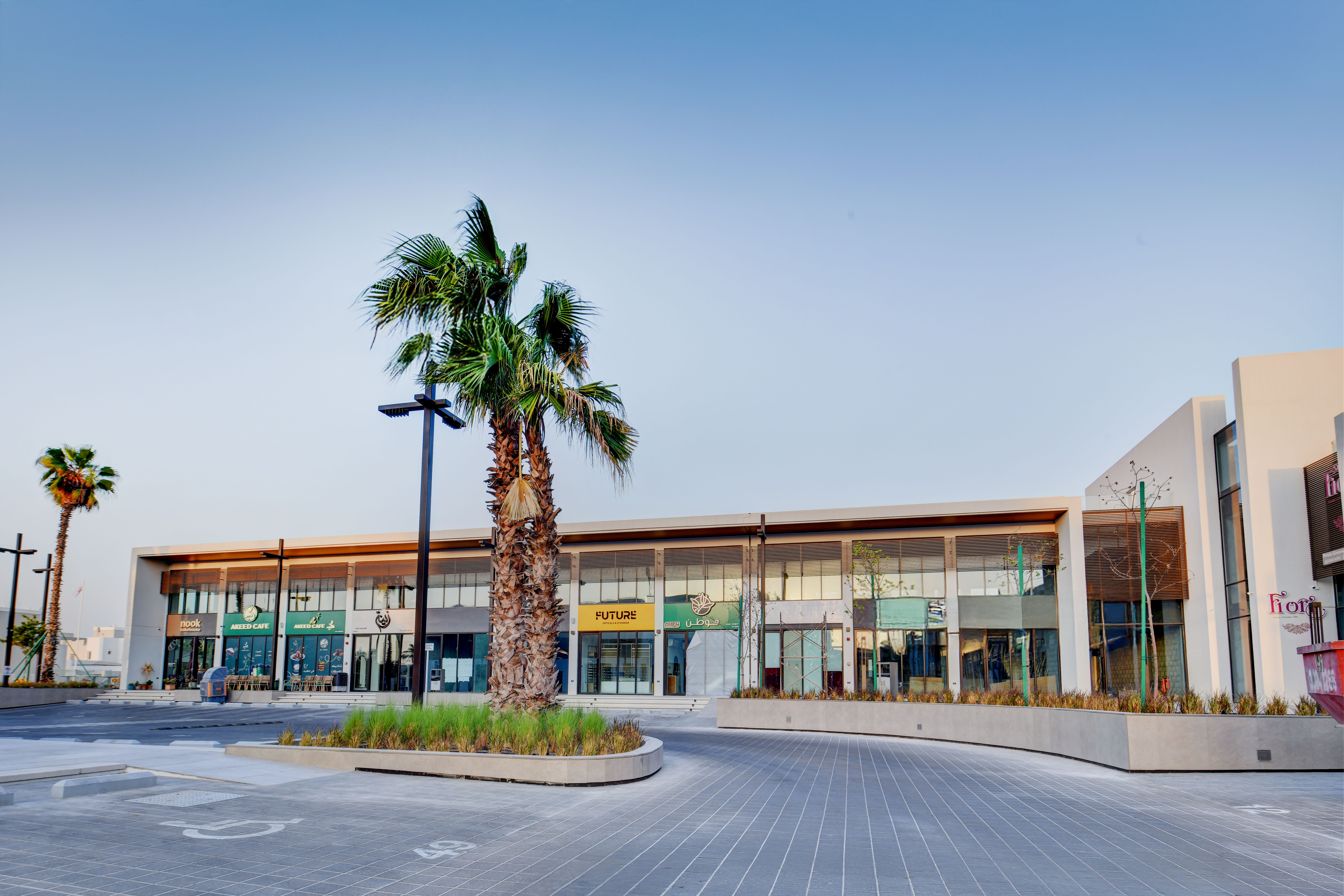 A modern two-story commercial building, East West Mall features large glass windows, palm trees, and empty parking spaces in front under a clear sky.