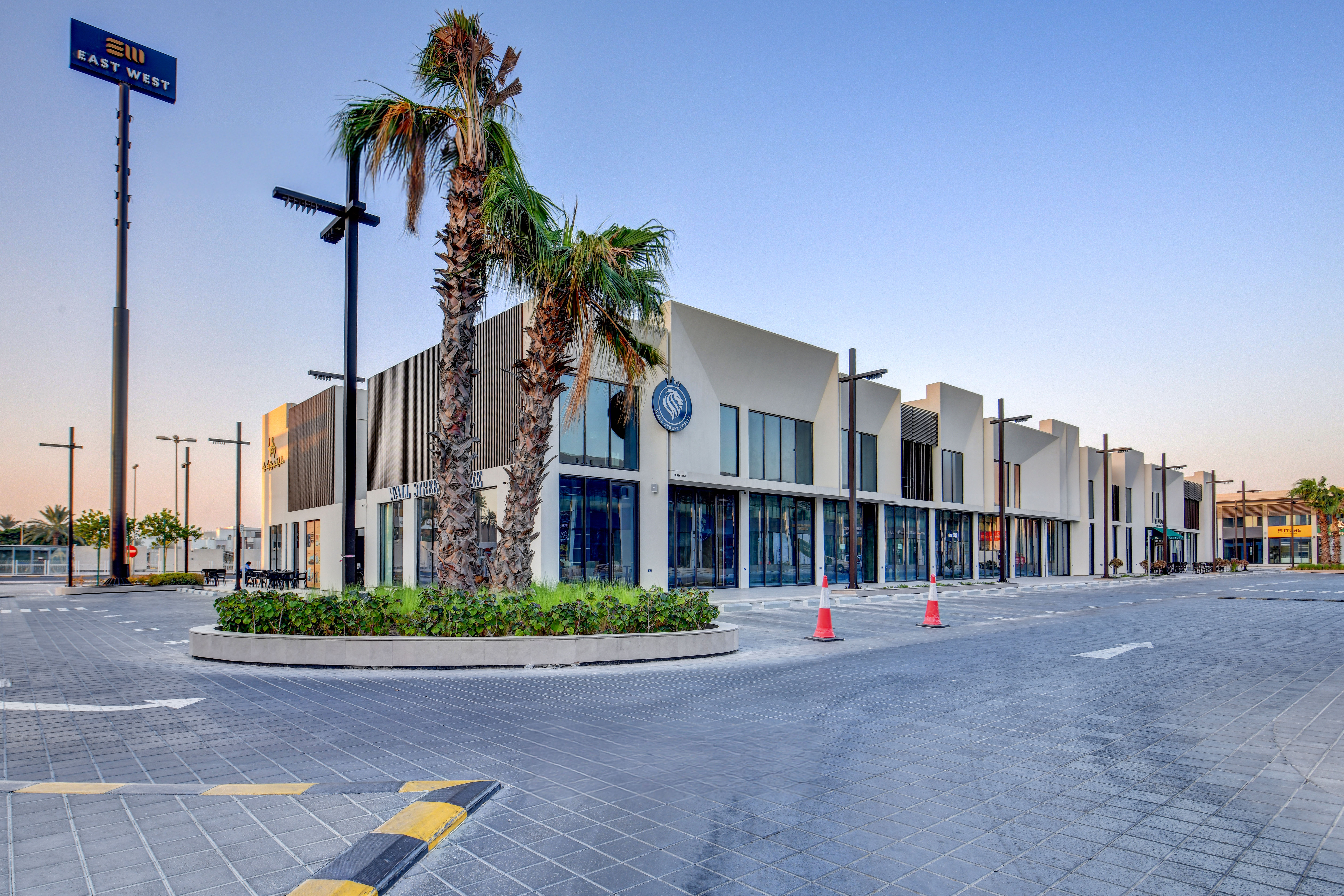 A modern commercial building, East West Mall, features large glass windows, palm trees, and an empty parking lot with traffic cones in front, all set under a clear sky.