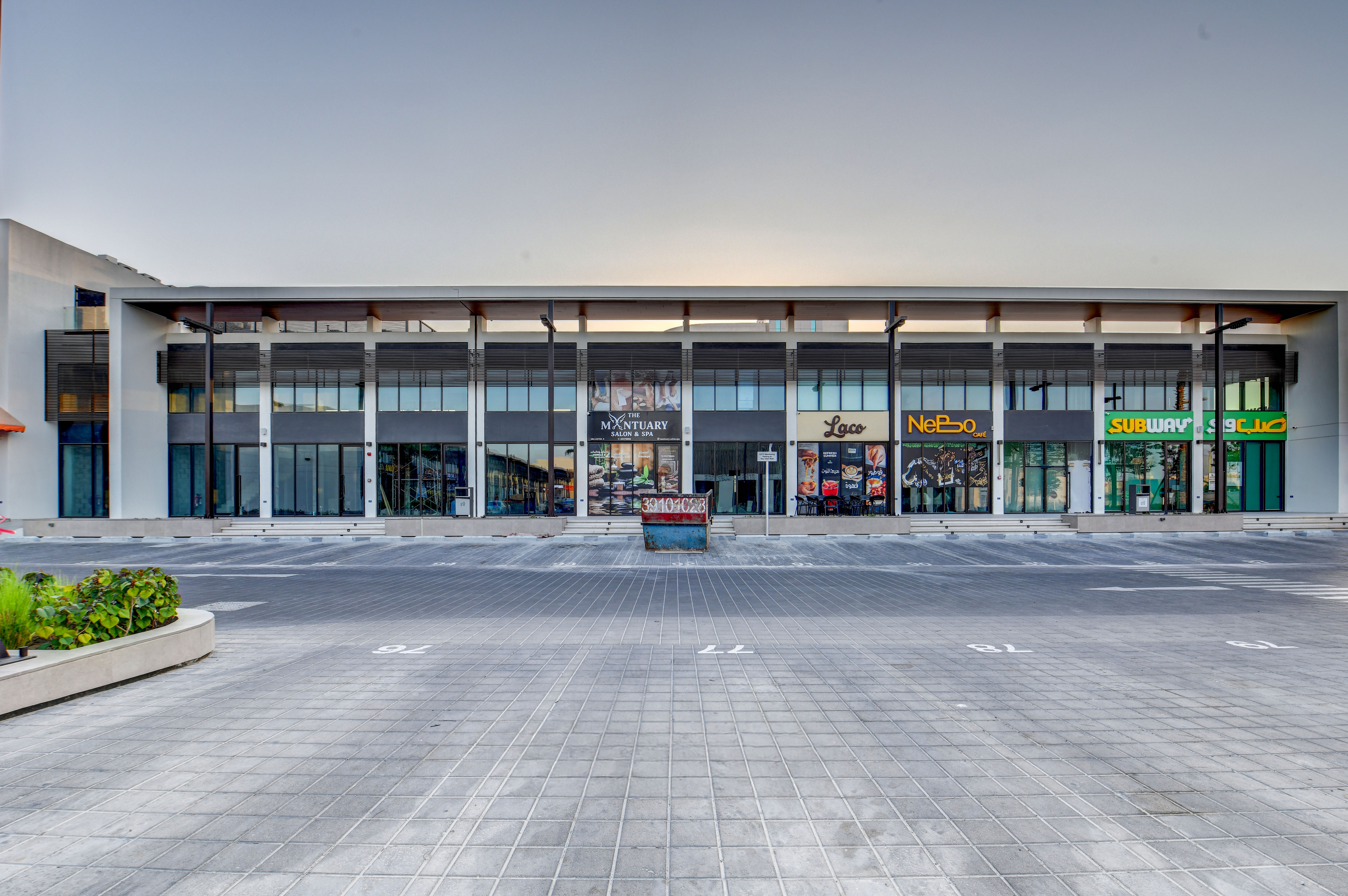 A modern commercial building with glass storefronts, various business signs, and an empty parking lot in front at East West Mall.