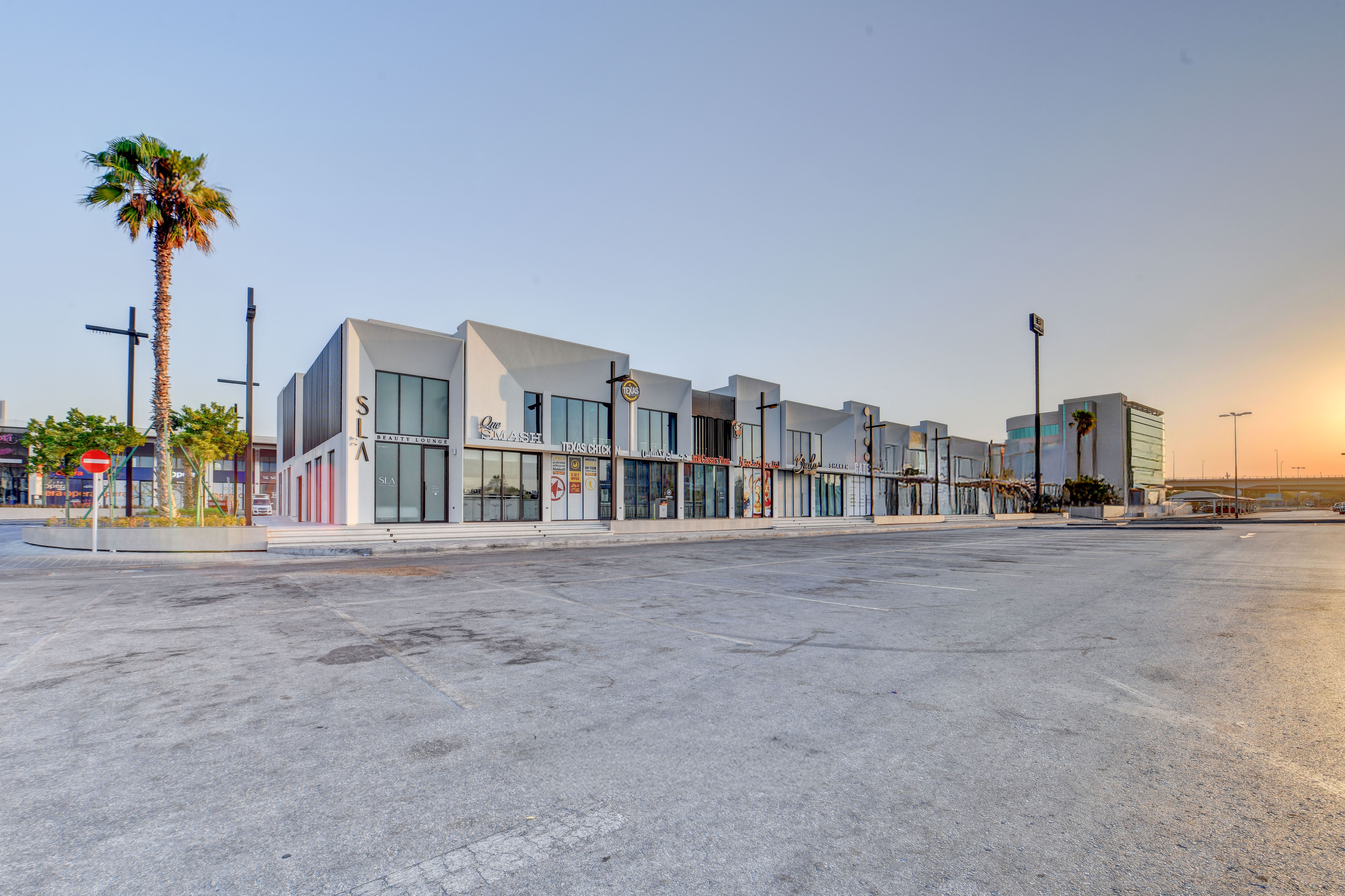 A modern strip of white, glass-fronted commercial buildings at East West Mall stands with a palm tree in front and an empty parking lot basking in the glow of sunrise.