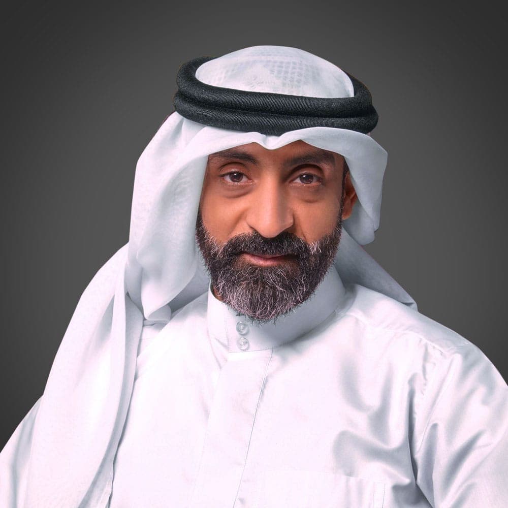 رجل يرتدي عباءة بيضاء وغترة باللونين الأبيض والأسود على خلفية داكنة عادية، وينظر مباشرة إلى الكاميرا.