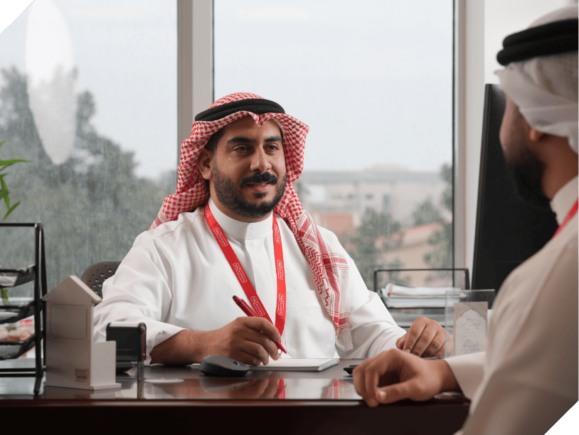 A man wearing traditional Middle Eastern attire and a red lanyard sits at a desk with a pen and notepad, discussing house me Bahrain careers with another person.