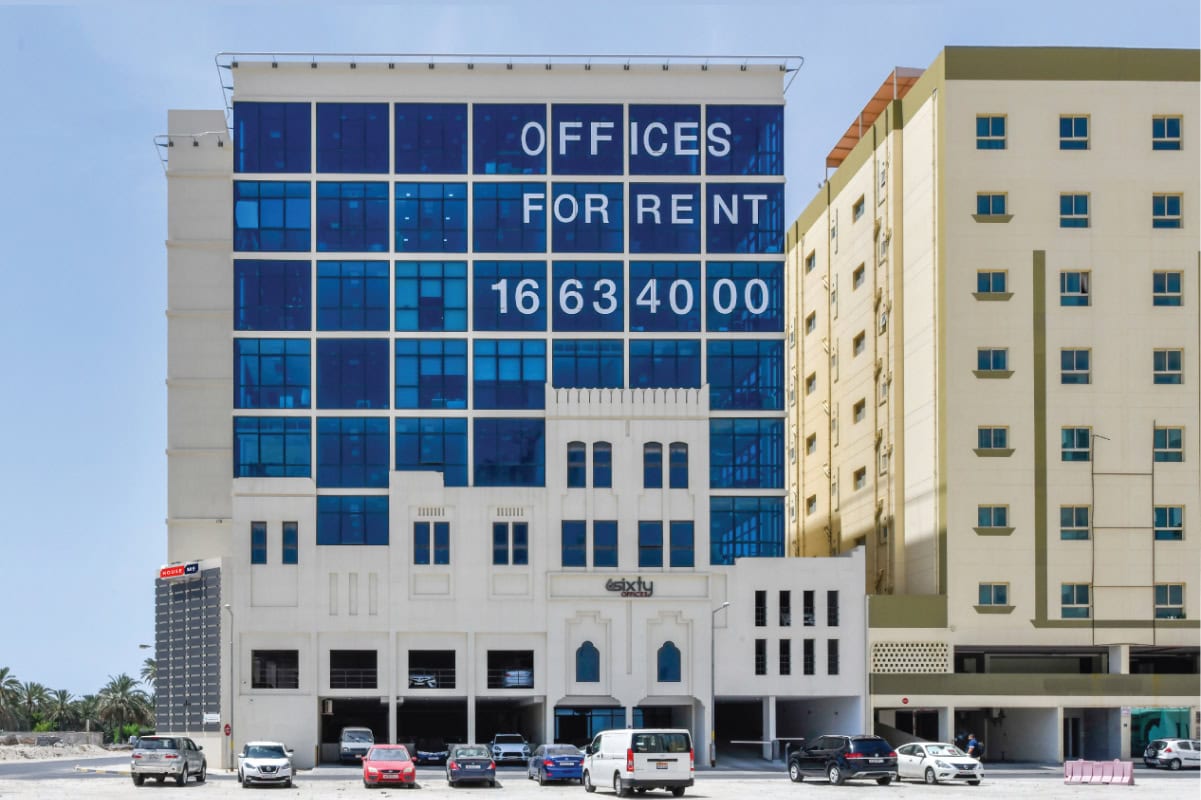 A modern office building with a large “Offices For Rent” sign and contact number displayed on its glass facade; several cars are parked in front.
