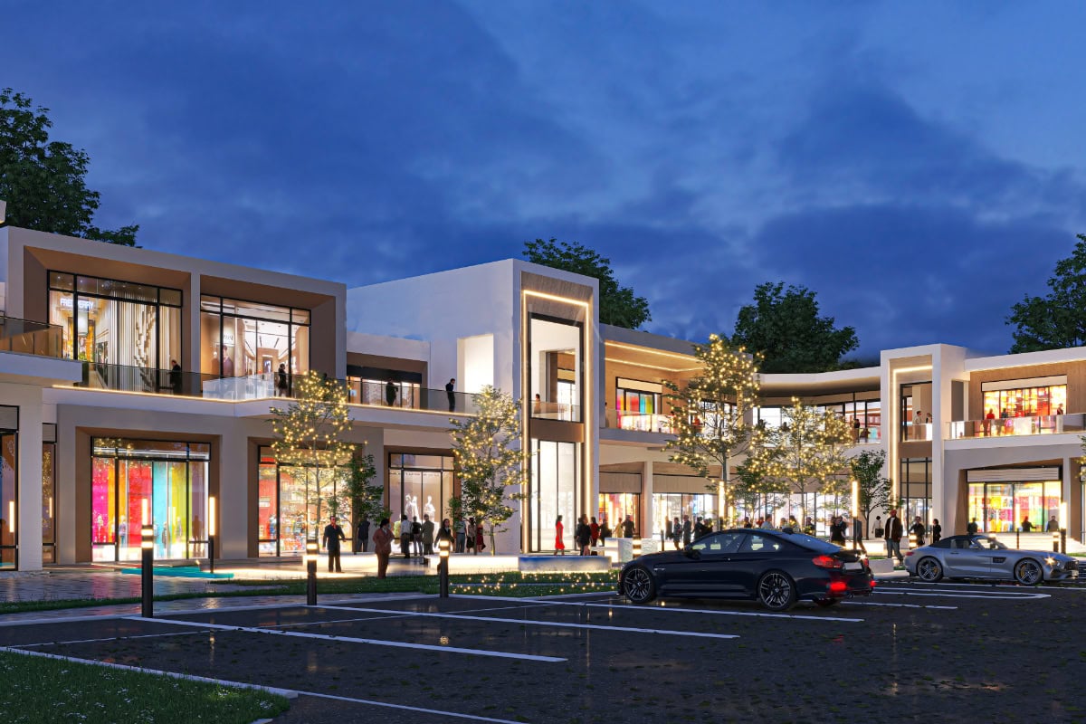 Modern shopping center at dusk with illuminated storefronts, large windows, parked cars in the foreground, and people walking near the entrance.