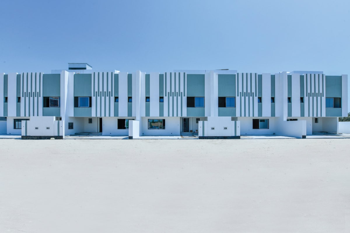 A modern white building with vertical blue and gray accents, featuring multiple windows and flat rooftops under a clear blue sky.