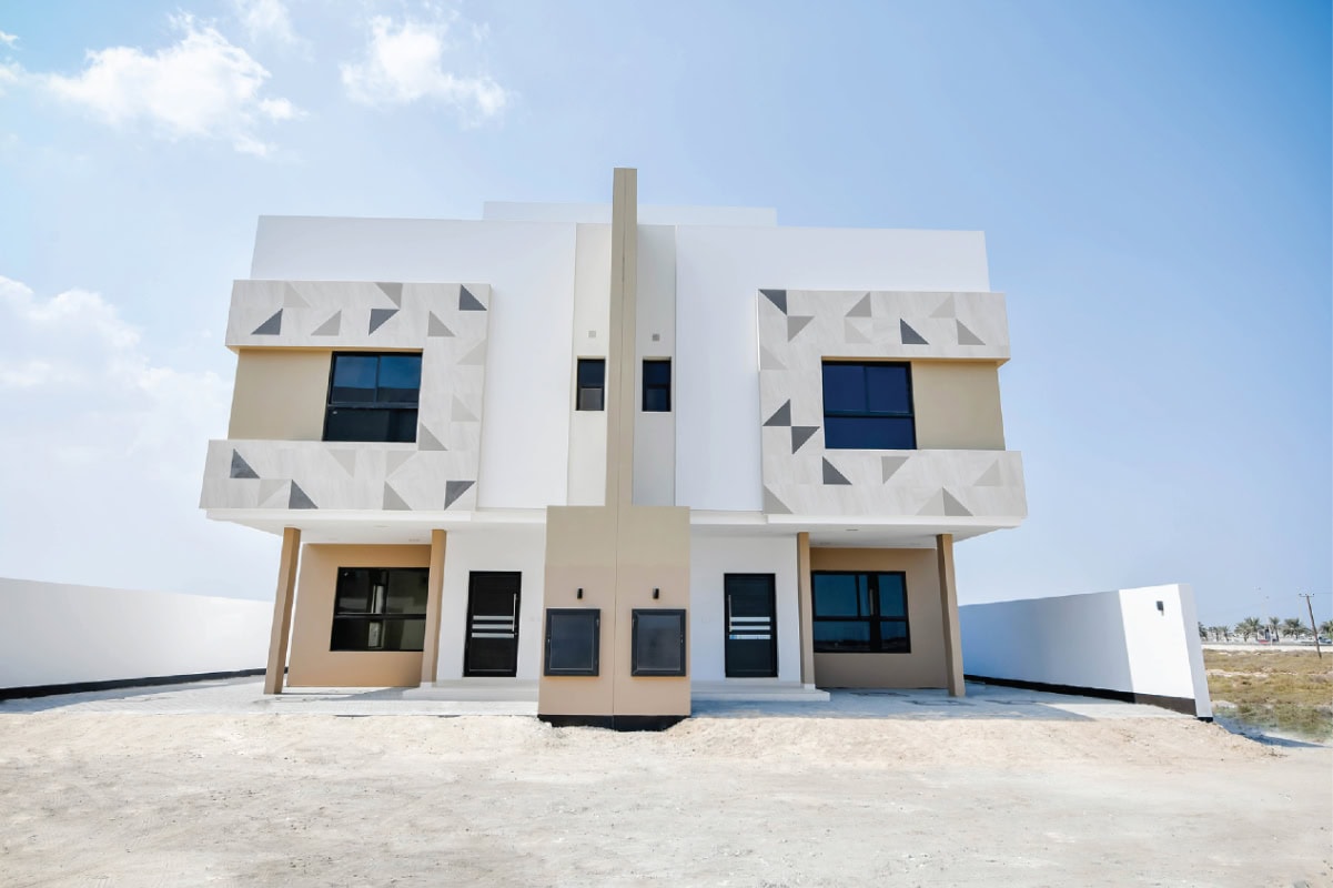 A modern, two-story duplex house with geometric patterns on the facade, large windows, and a minimalistic exterior, situated on a sandy lot under a clear sky.