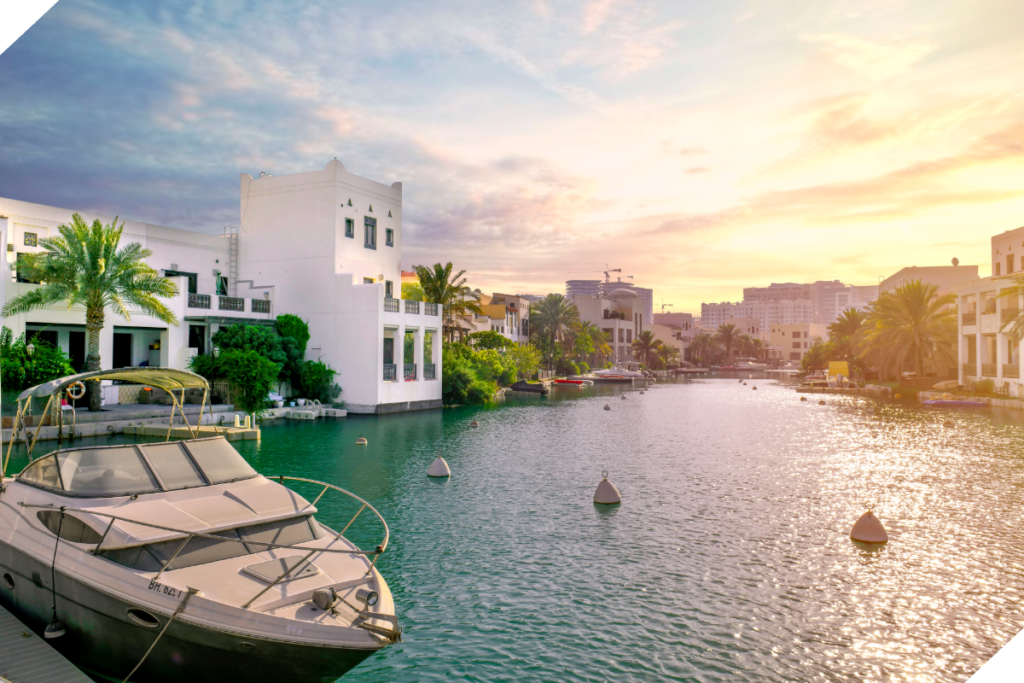 A white building and palm trees line a canal with a docked boat in the foreground, under a colorful sunset sky.
