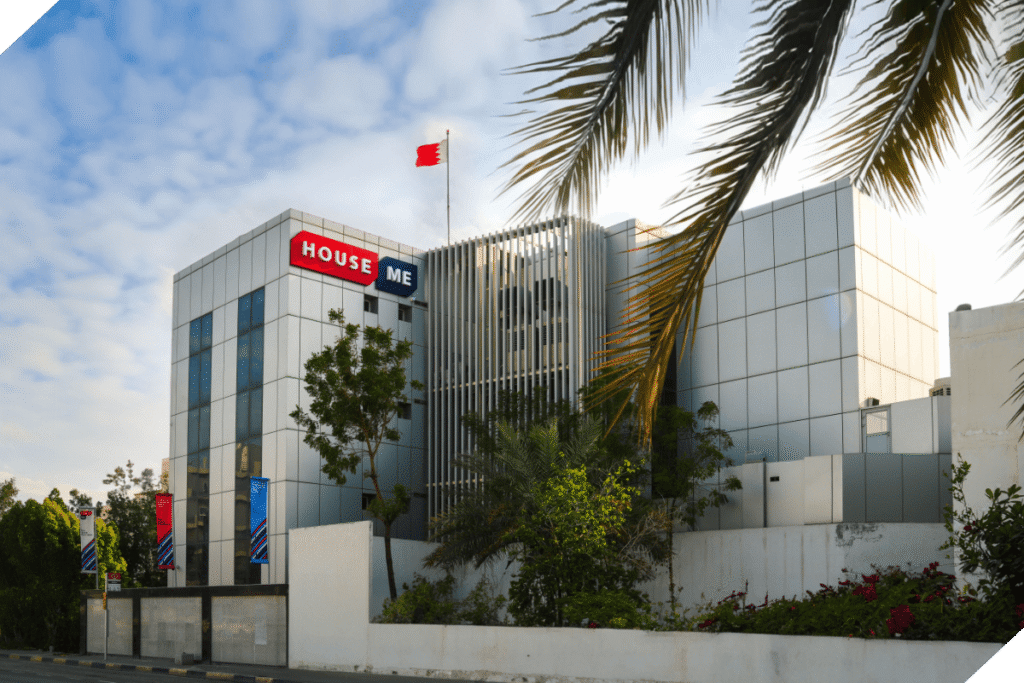 Modern office building with reflective glass exterior, a "HOUSE ME" sign, and a flag on the roof, surrounded by palm trees and greenery under a partly cloudy sky.