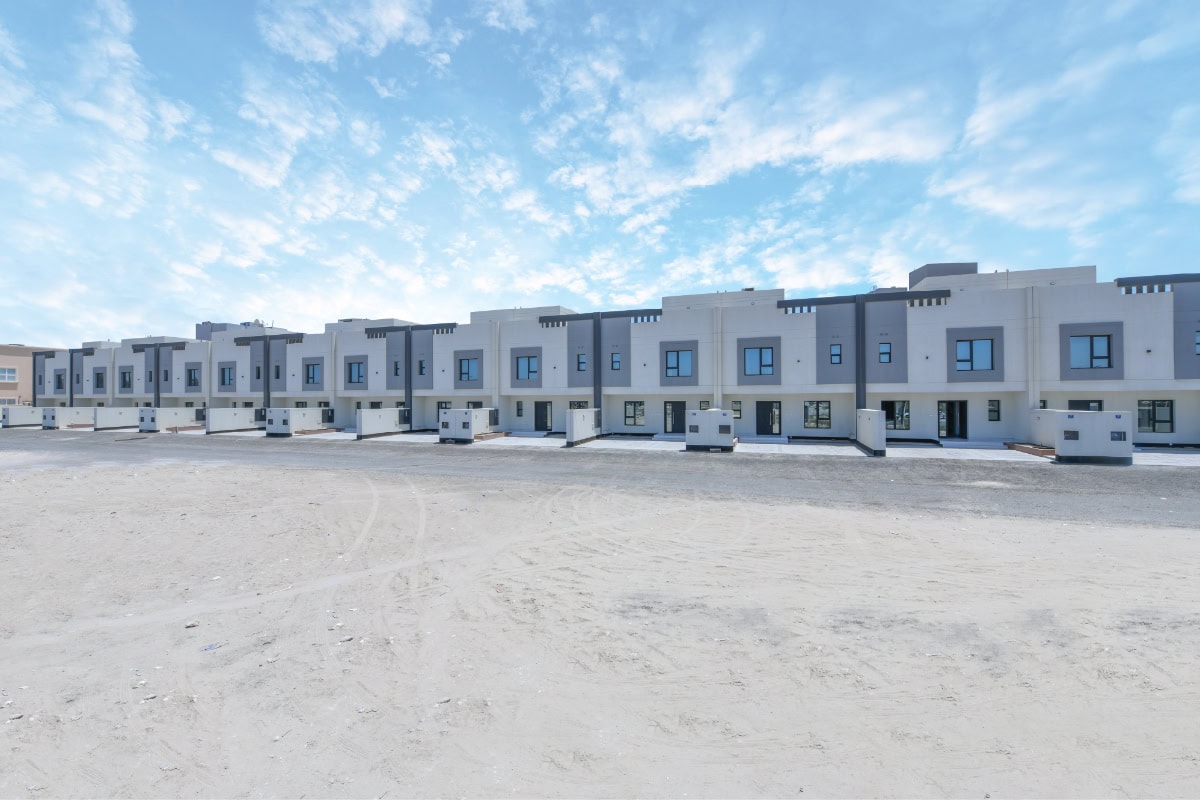 Row of modern two-story townhouses with flat roofs, uniform design, and driveways, set against a blue sky with scattered clouds and a barren foreground.