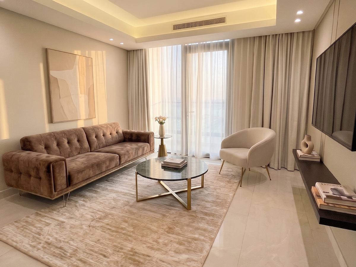 Modern living room by Memaar Amwaj with a brown sofa, round glass coffee table, cream accent chair, abstract wall art, floor-to-ceiling curtains, and neutral decor.