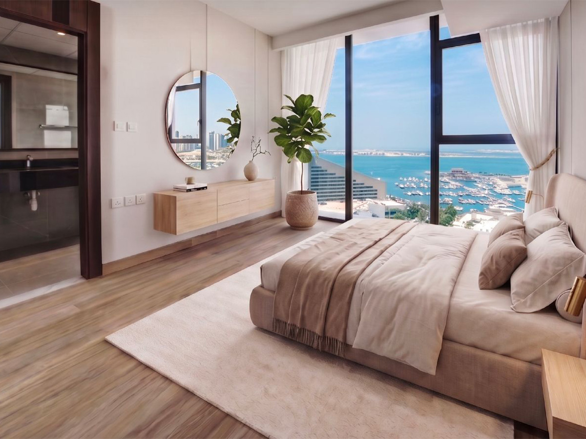 Modern bedroom at Memaar Amwaj with large windows, neutral décor, a potted plant, wall-mounted mirror, and stunning views of the ocean, marina, and city skyline.