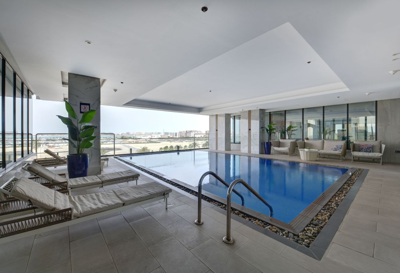 Indoor pool with metal ladder, surrounded by lounge chairs and potted plants, next to large windows overlooking the Memaar Amwaj cityscape.