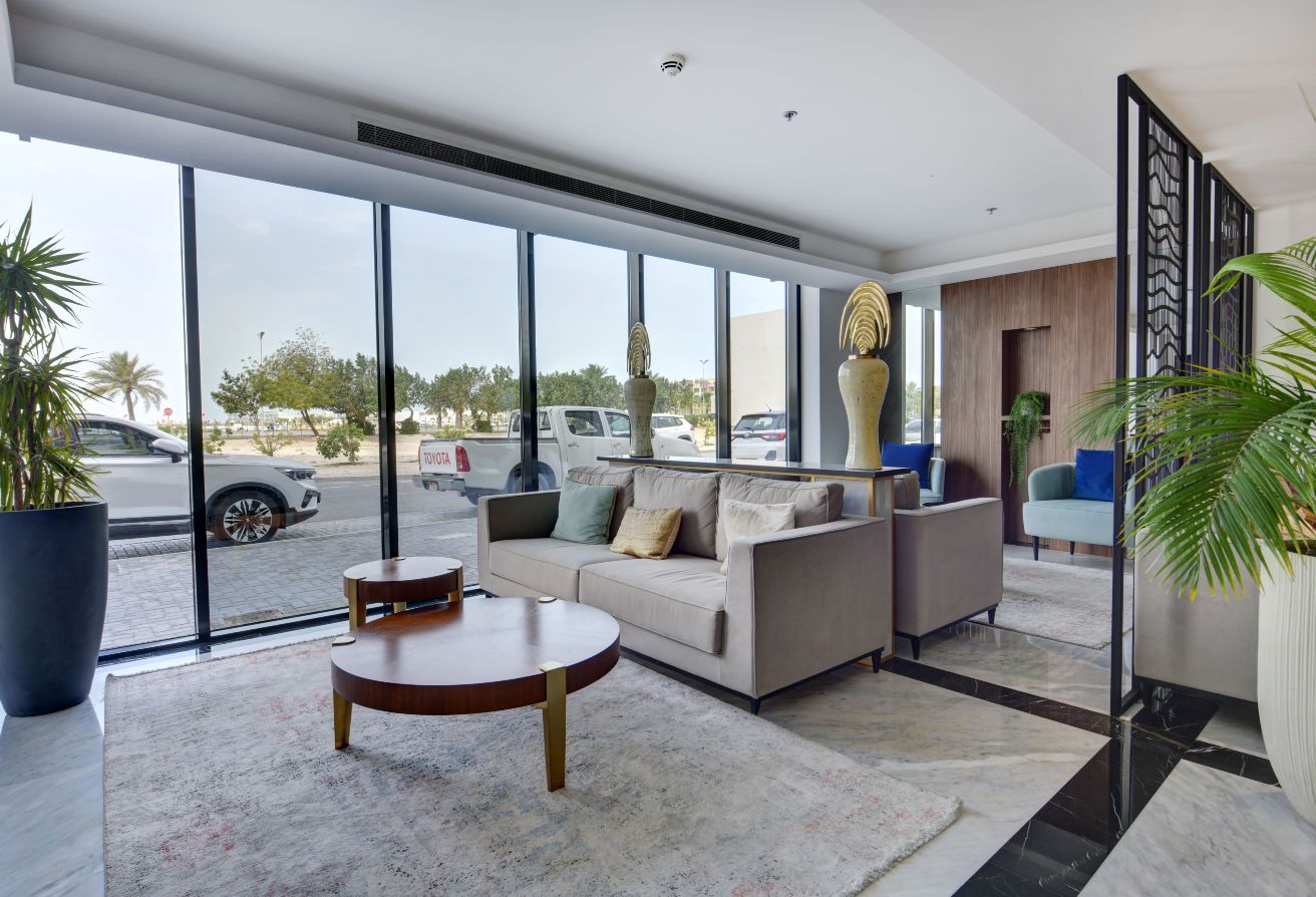 Modern hotel lobby at Memaar Amwaj featuring a beige sofa, round coffee tables, large windows, indoor plants, and parked cars visible outside.