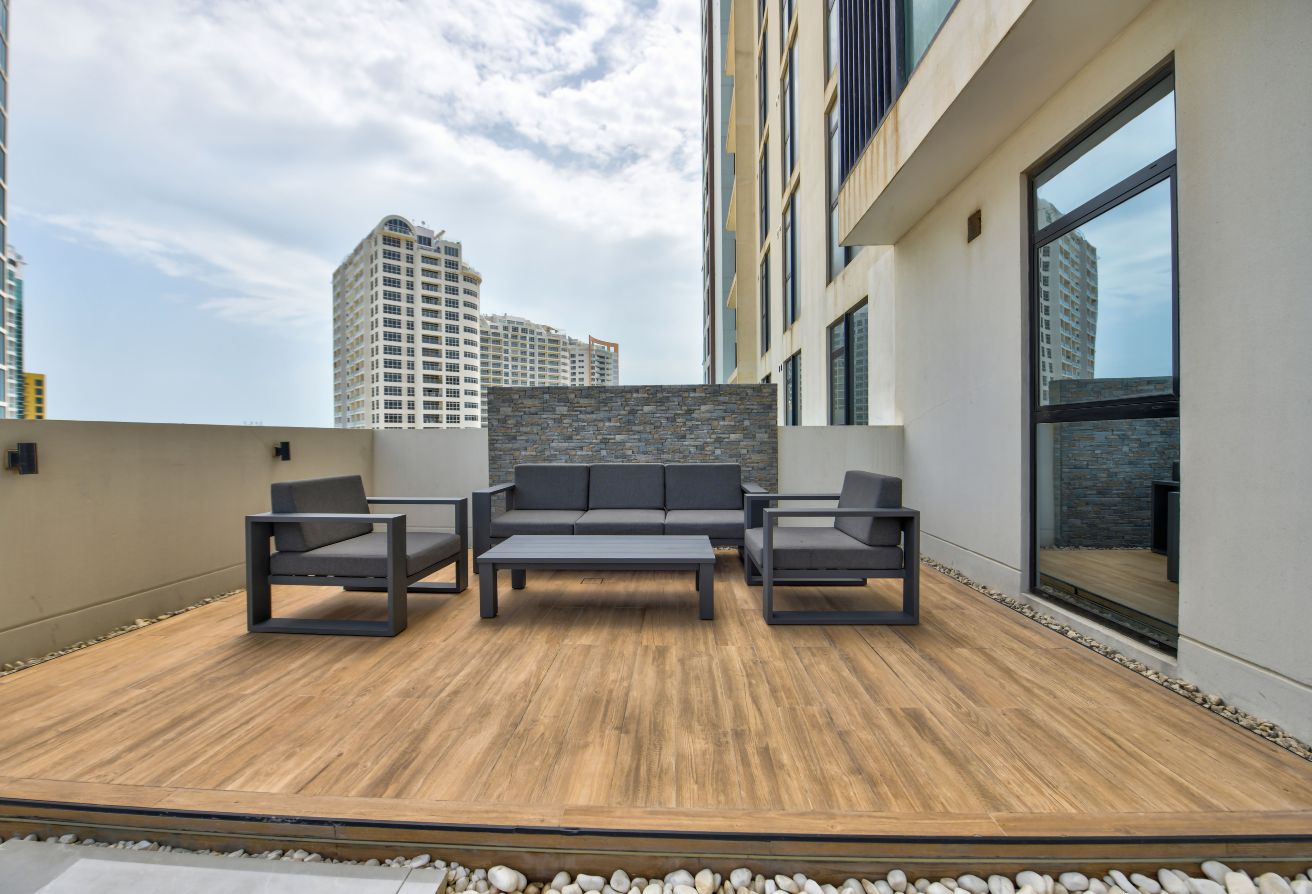 Modern outdoor patio with gray furniture on a wooden deck by Memaar, surrounded by tall buildings under a partly cloudy sky—an urban oasis inspired by Amwaj living.