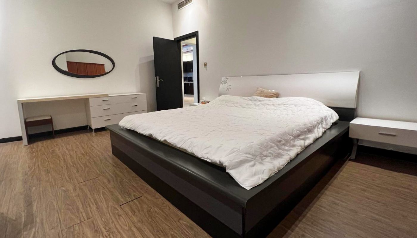 Modern bedroom with a low black platform bed, white bedding, wooden floor, a desk with a mirror, and a nightstand, all against plain white walls.