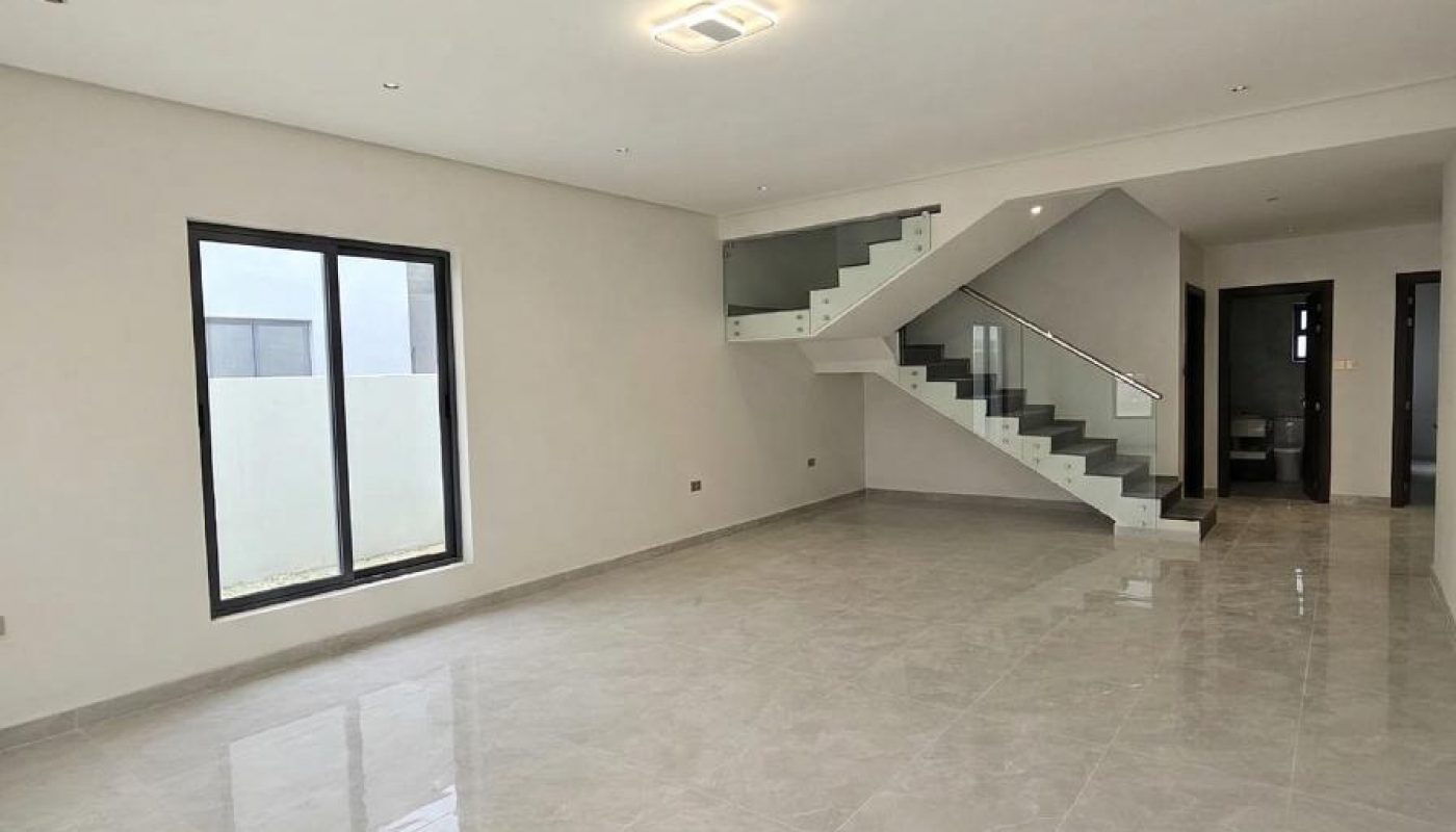 Spacious, empty living room with tiled floors, white walls, large window, and a modern staircase with glass railing leading to an upper floor.