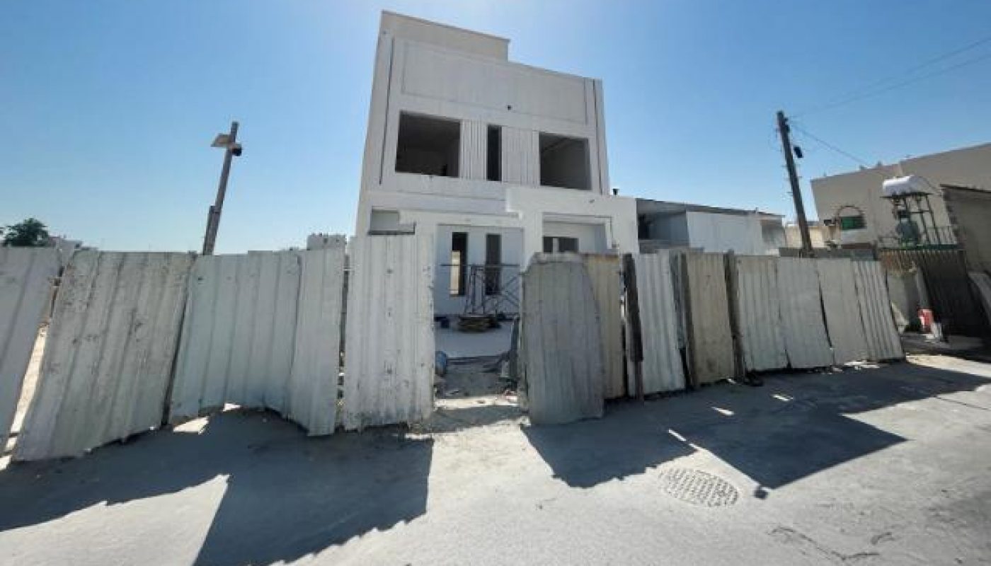 A three-story white building under construction, surrounded by a corrugated metal fence, with a bright blue sky and sunlight overhead.