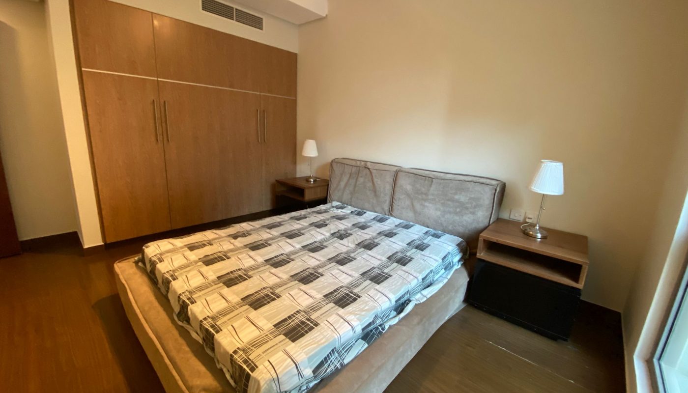 A bedroom with a double bed featuring a checkered bedspread, two bedside tables with lamps, a large wooden wardrobe, and wooden flooring.