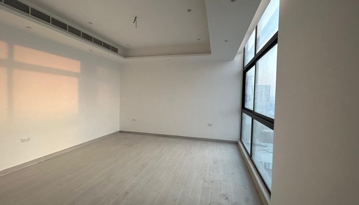 Empty room with light gray wood flooring, white walls, large floor-to-ceiling windows on the right, and recessed ceiling lights; electrical outlet visible on back wall.