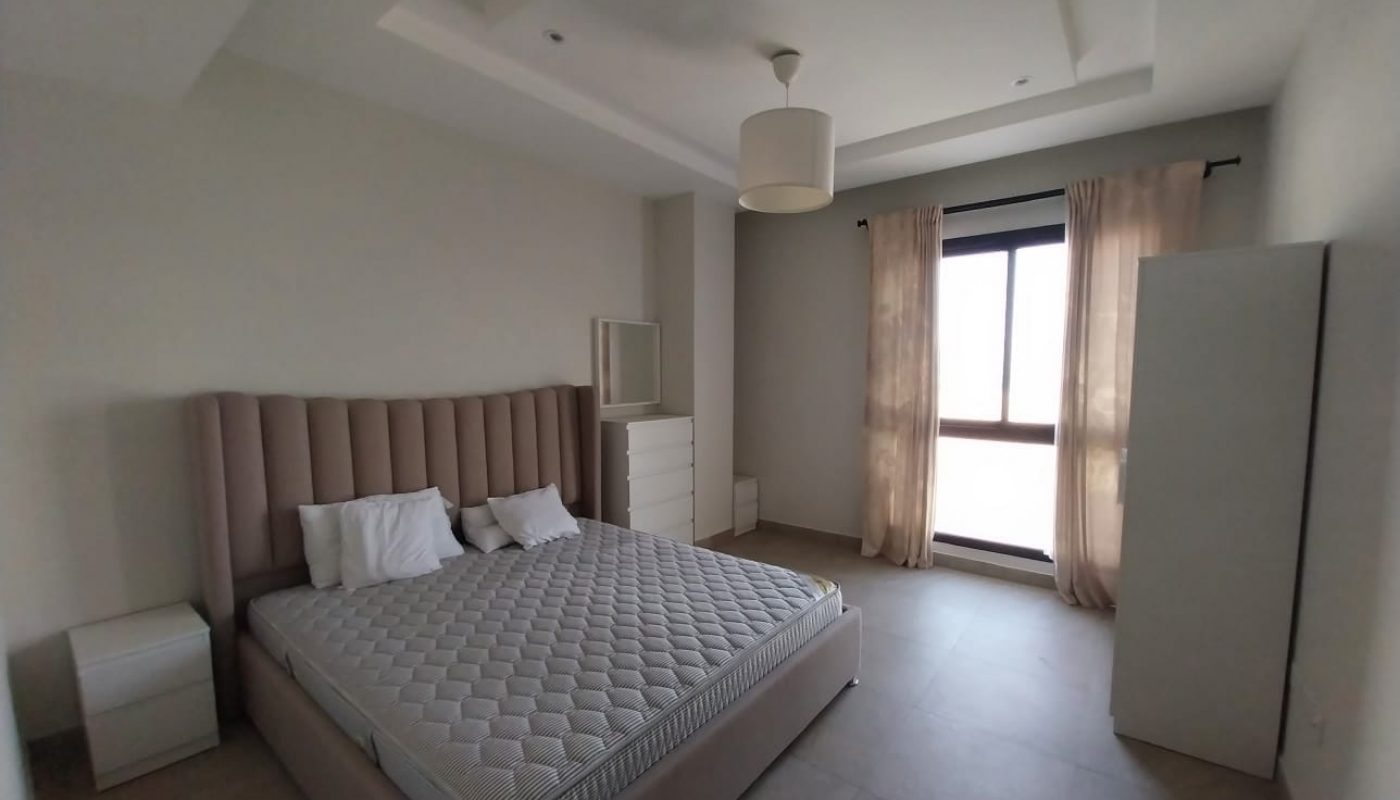 A minimalist bedroom in a flat for rent featuring a large bed with a padded headboard, two side tables, a chest of drawers with a mirror, and a window with curtains allowing natural light to enter the room.