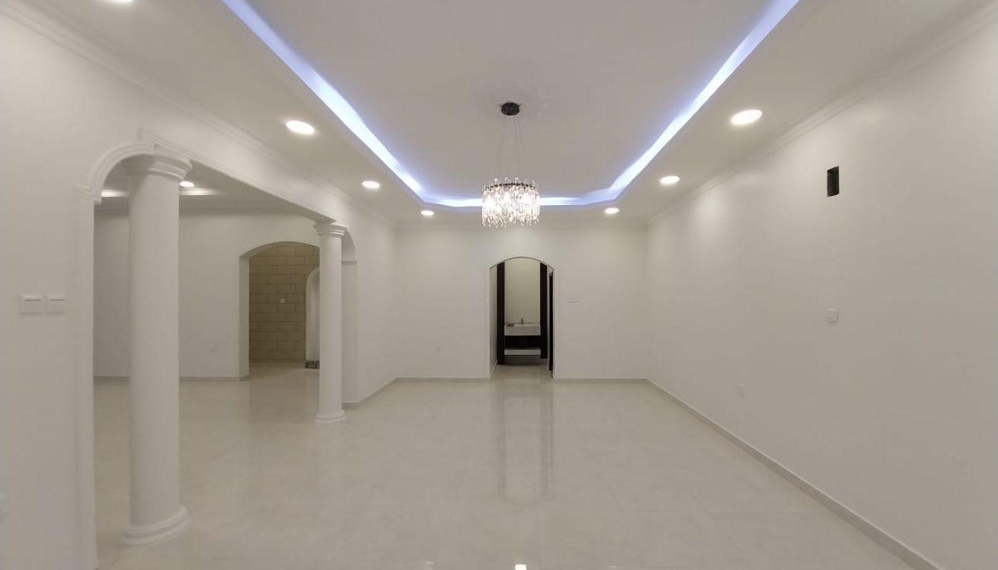 Spacious, empty white room with glossy tiled floor, columns, recessed ceiling lights, blue accent lighting, and a chandelier in the center.