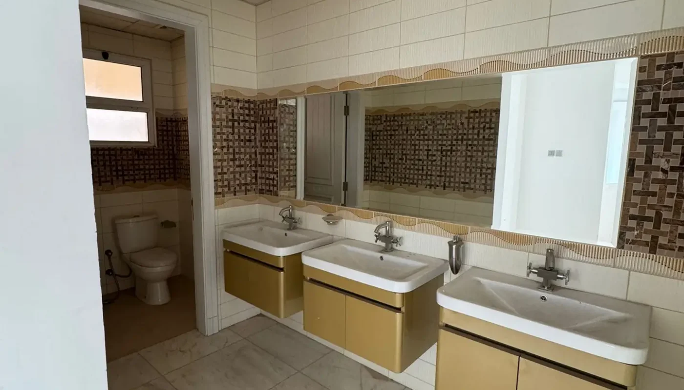 A bathroom with two sinks, a large mirror, tiled walls, and an adjacent room containing a toilet. Marble flooring and overhead ceiling panels are visible.