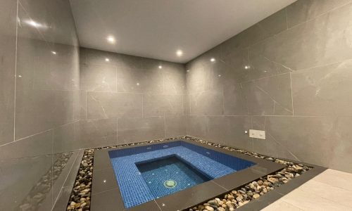 A minimalist indoor spa room in this AMWAJ flat for sale features a small, rectangular blue-tiled hot tub surrounded by gray tiles and decorative pebbles.